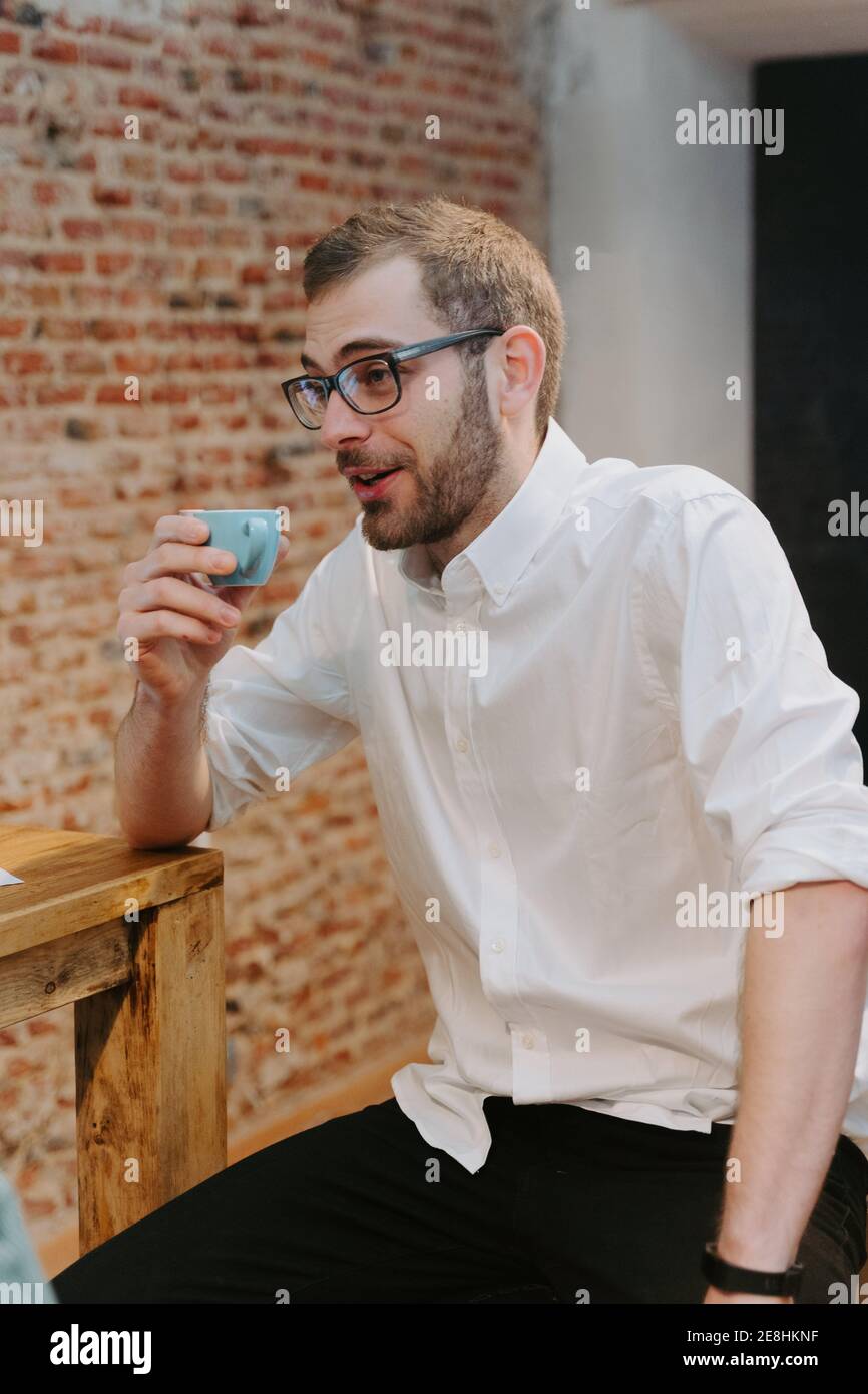 Barista maschile in camicia bianca e con piccola tazza di espresso seduto al bancone in legno nel caffè e guardando lontano Foto Stock