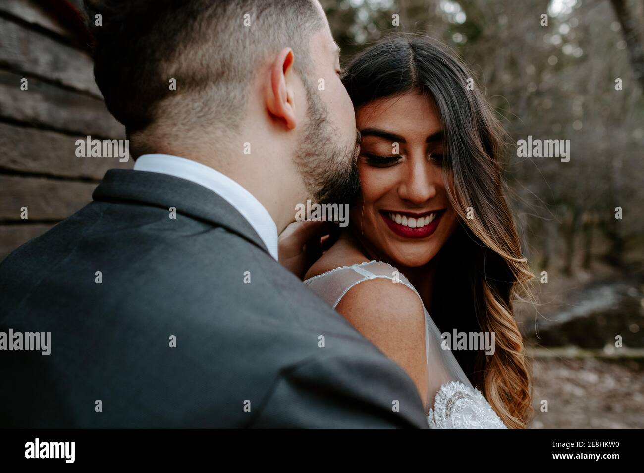 Vista laterale dello sposo giovane in elegante tuta baciante sorridente sposa etnica mentre si levano in piedi nella natura il giorno del matrimonio Foto Stock