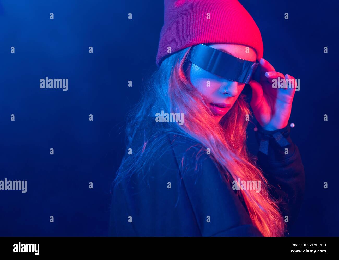 Giovane donna millennial trendy con lunghi capelli biondi in futuristico occhiali da sole che regolano il cappello in piedi in camera oscura con neon illuminazione Foto Stock