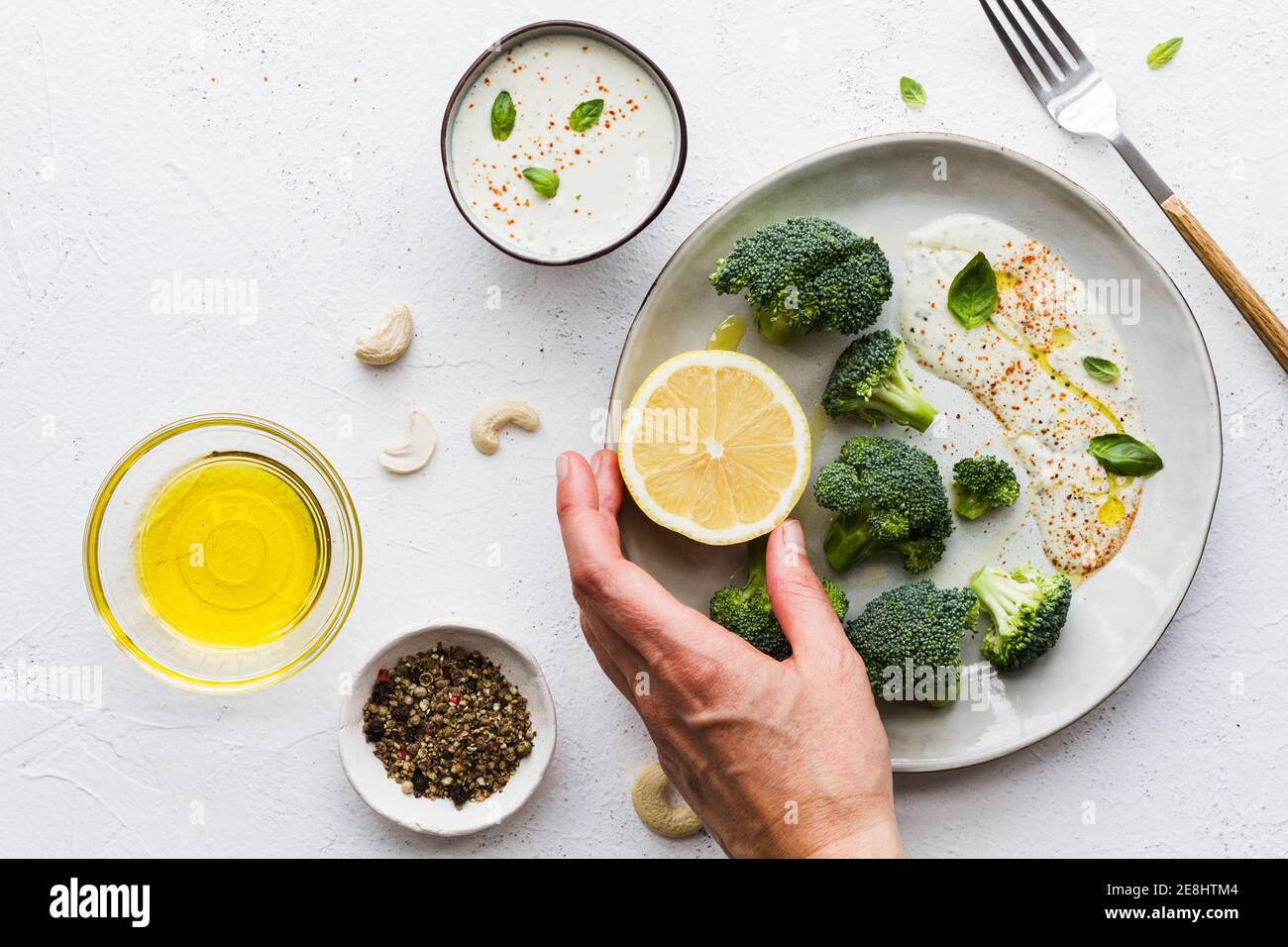 Vista dall'alto delle mani di una persona non riconoscibile che mangia gustosi broccoli con limone fresco e salsa vicino a ciotole con olio d'oliva e composto di spezie sul tavolo Foto Stock