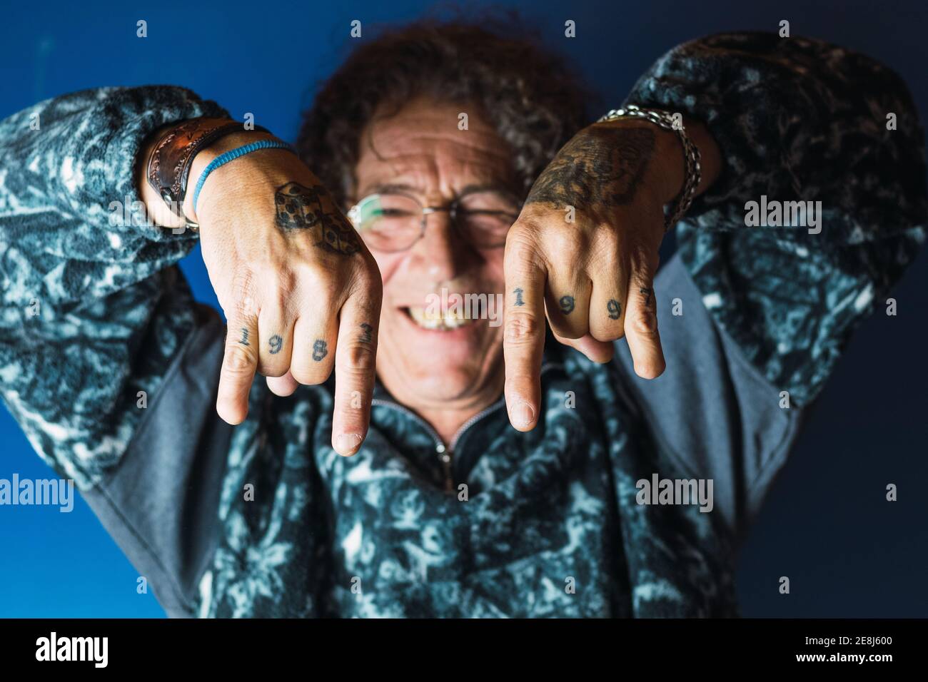 Allegro maschio di mezza età con capelli ricci e tatuaggi su mani senza un pollice in outfit trendy e occhiali sorridenti e mostrando movimento rock n roll Foto Stock