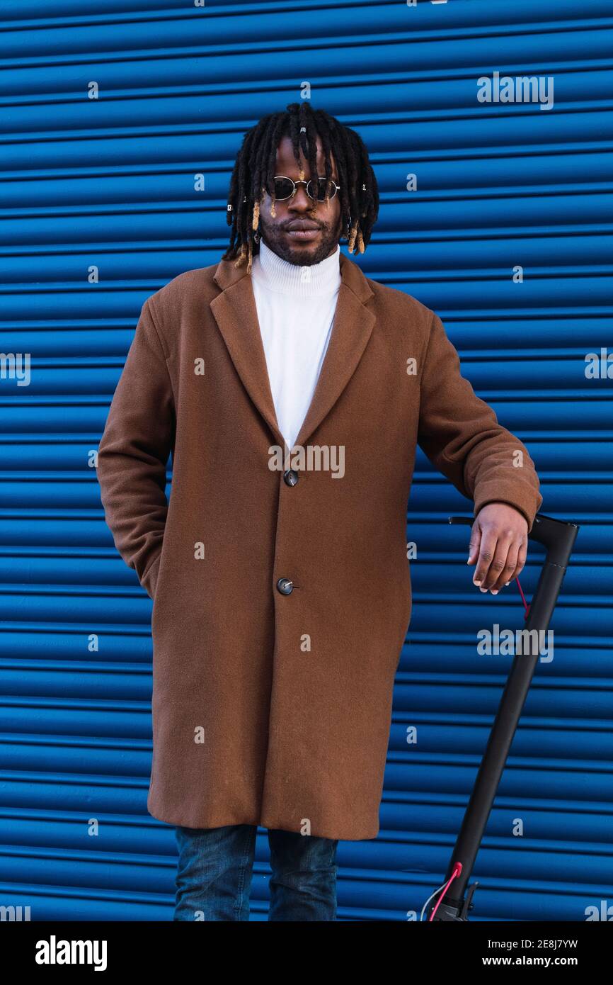 Uomo sicuro dell'hipster afroamericano adulto con le trecce di afro che  indossano cappotto elegante e occhiali da sole in piedi vicino scooter  elettrico contro il blu a strisce Foto stock - Alamy