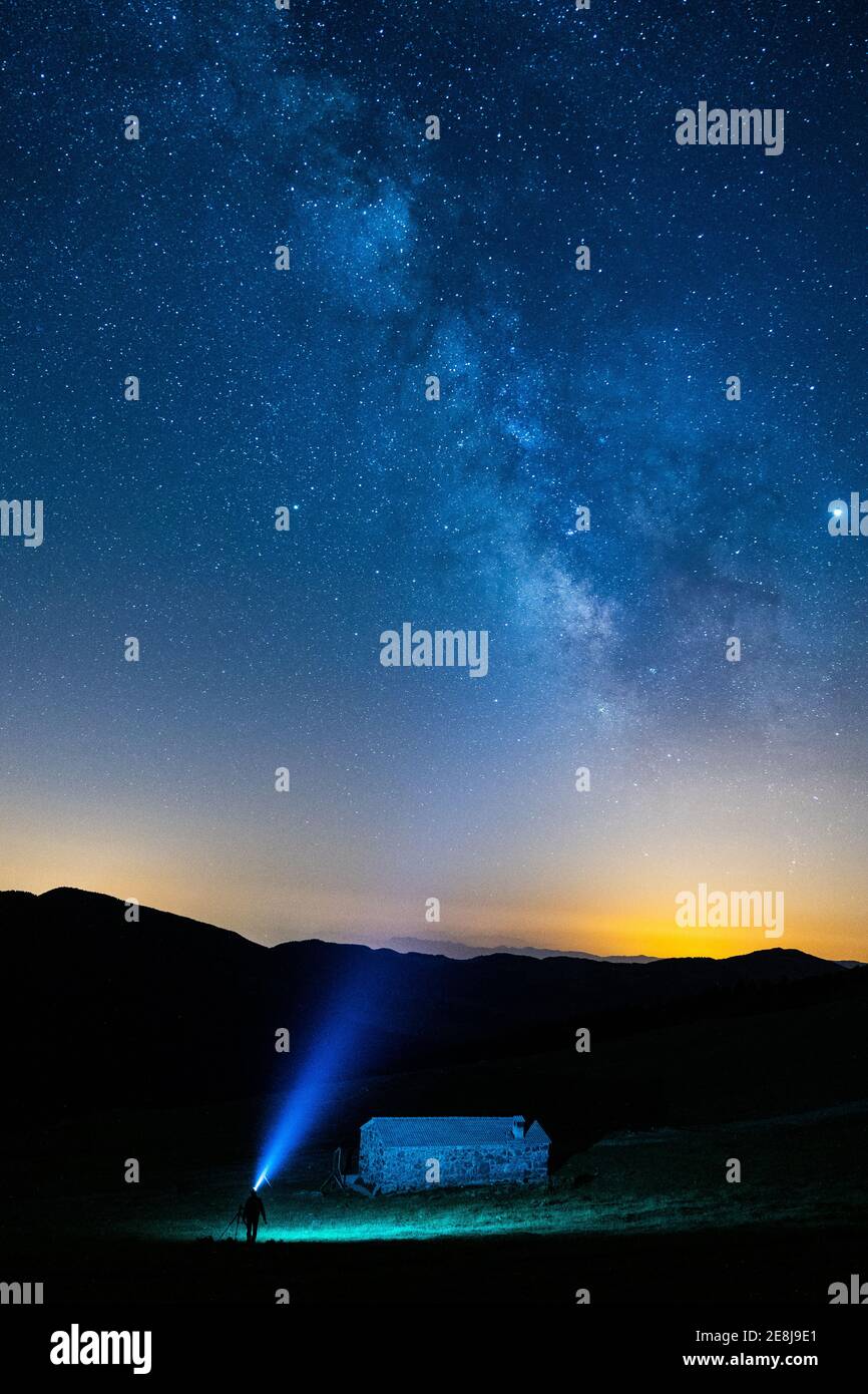 Silhouette viaggiatore anonima con luce blu in piedi vicino montagna sotto cielo colorato con stelle di notte Foto Stock