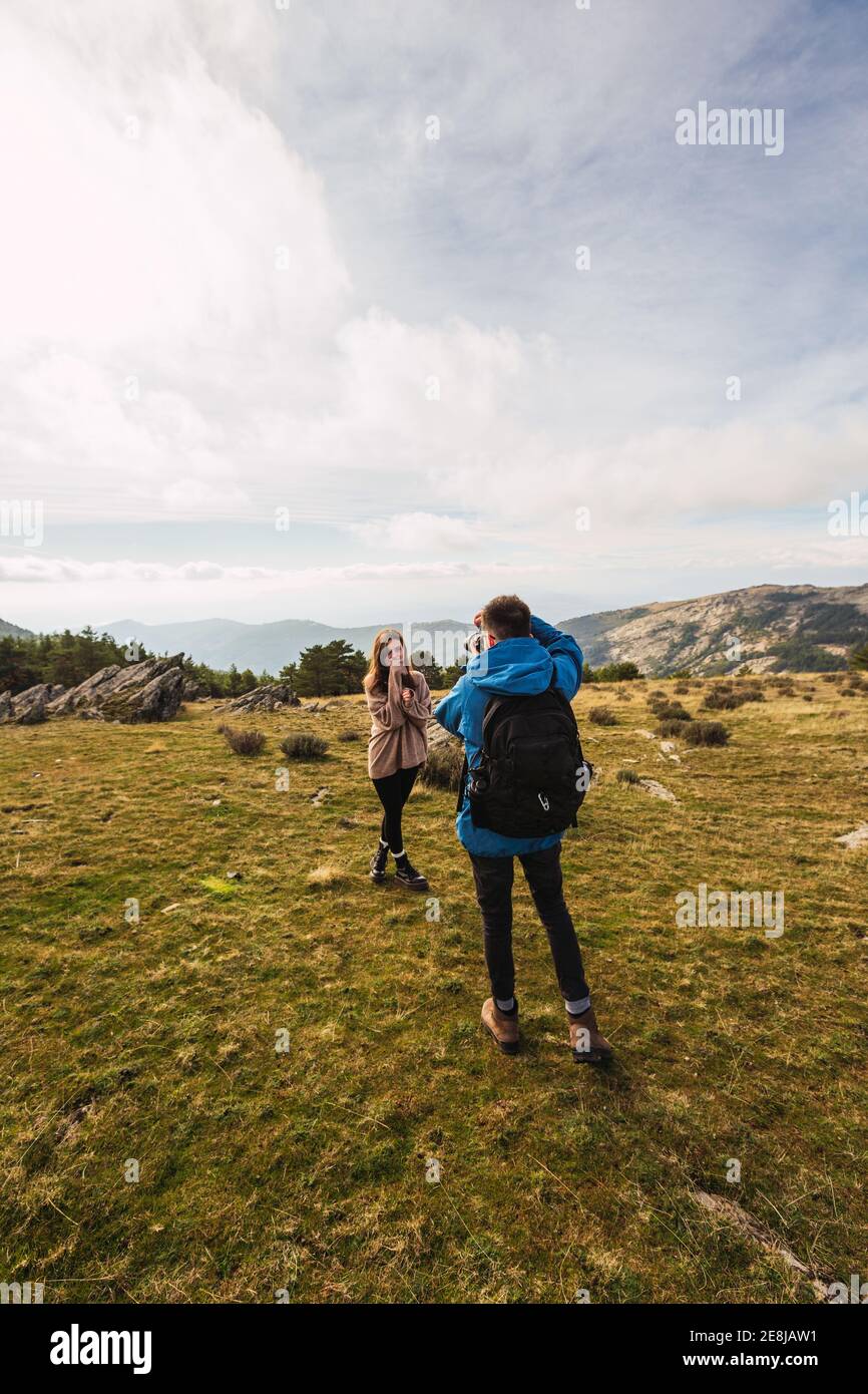 Uomo irriconoscibile trekking scattare foto di partner femminile sulla fotocamera mentre si è in piedi sul prato contro il monte sotto il cielo nuvoloso Foto Stock