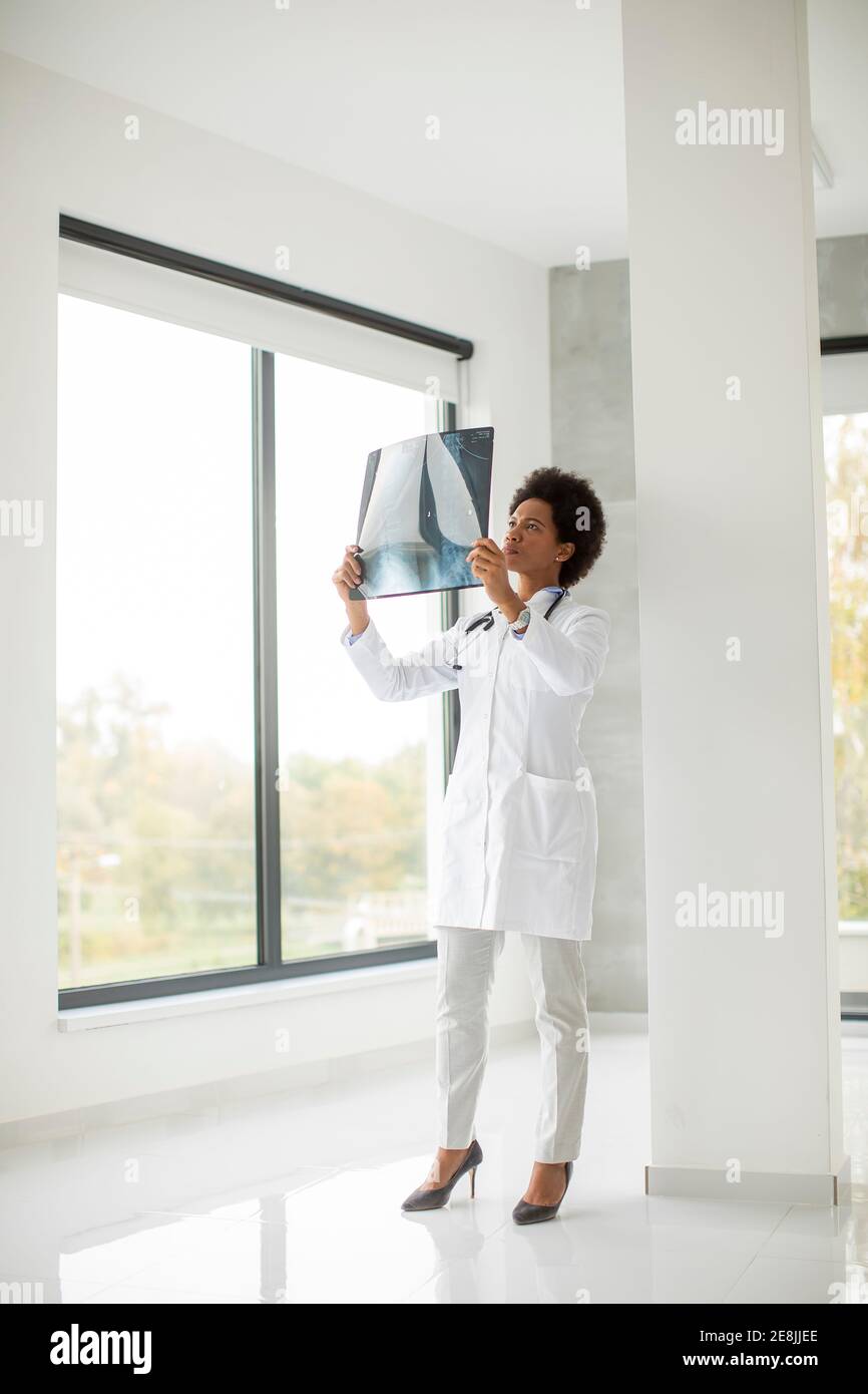Donna Africana americana medico indossando camice bianco con stetoscopio in piedi dalla finestra in ufficio e guardando un'immagine radiografica Foto Stock
