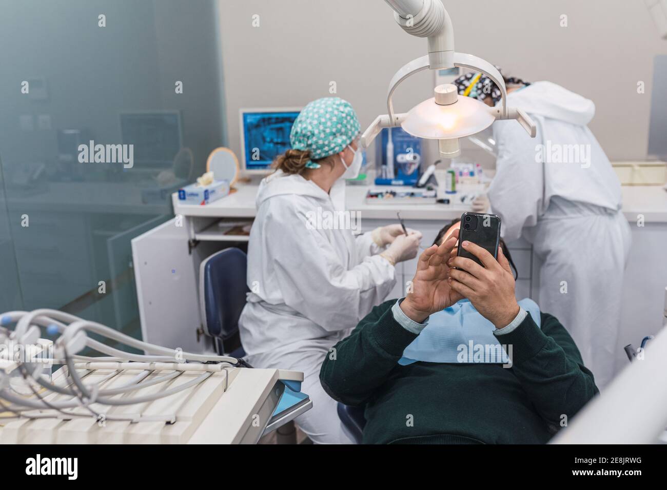 Paziente che utilizza uno smartphone in studio dentistico. Due dottoresse che preparano la procedura. Concetto di igiene e salute dentale Foto Stock