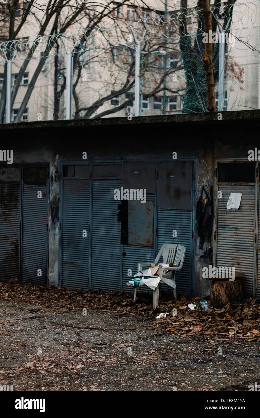 Sedia grigia bianca rotta di fronte al vecchio blu porta del garage arrugginita con recinzione in filo spinato sopra Foto Stock