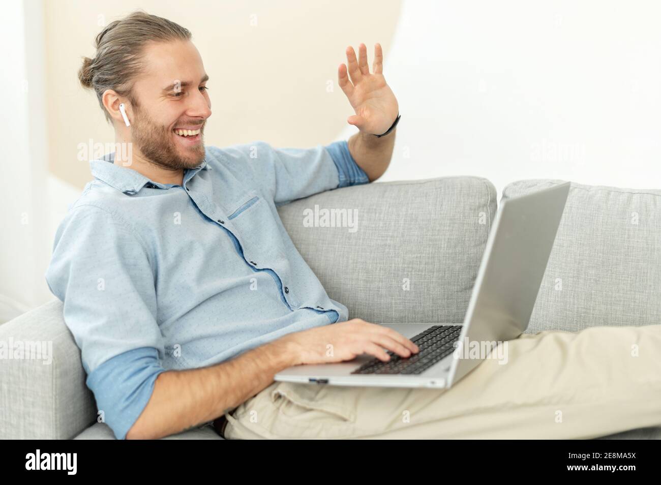 Un hipster sorridente è seduto sul comodo divano del soggiorno, rilassandosi e parlando con un amico o con la famiglia, sventolando al computer portatile Foto Stock