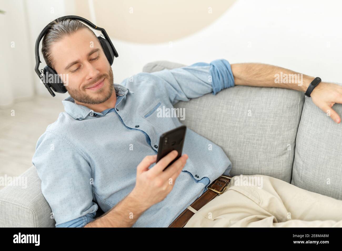 Attraente giovane hipster sopportato millenario indossando le cuffie, distendersi e distendersi sul divano comodo, ascoltare la musica, chattare con Foto Stock