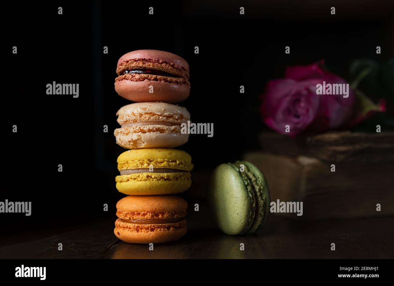 Pila di deliziosi macaron assortiti di colori diversi, un vecchio libro e una rosa su uno sfondo scuro Foto Stock
