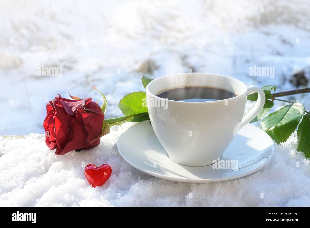 Caffè caldo in una tazza bianca servito all'aperto nella neve, decorato con una rosa rossa e un piccolo cuore dal vetro in una giornata di sole inverno, concetto di amore per Foto Stock