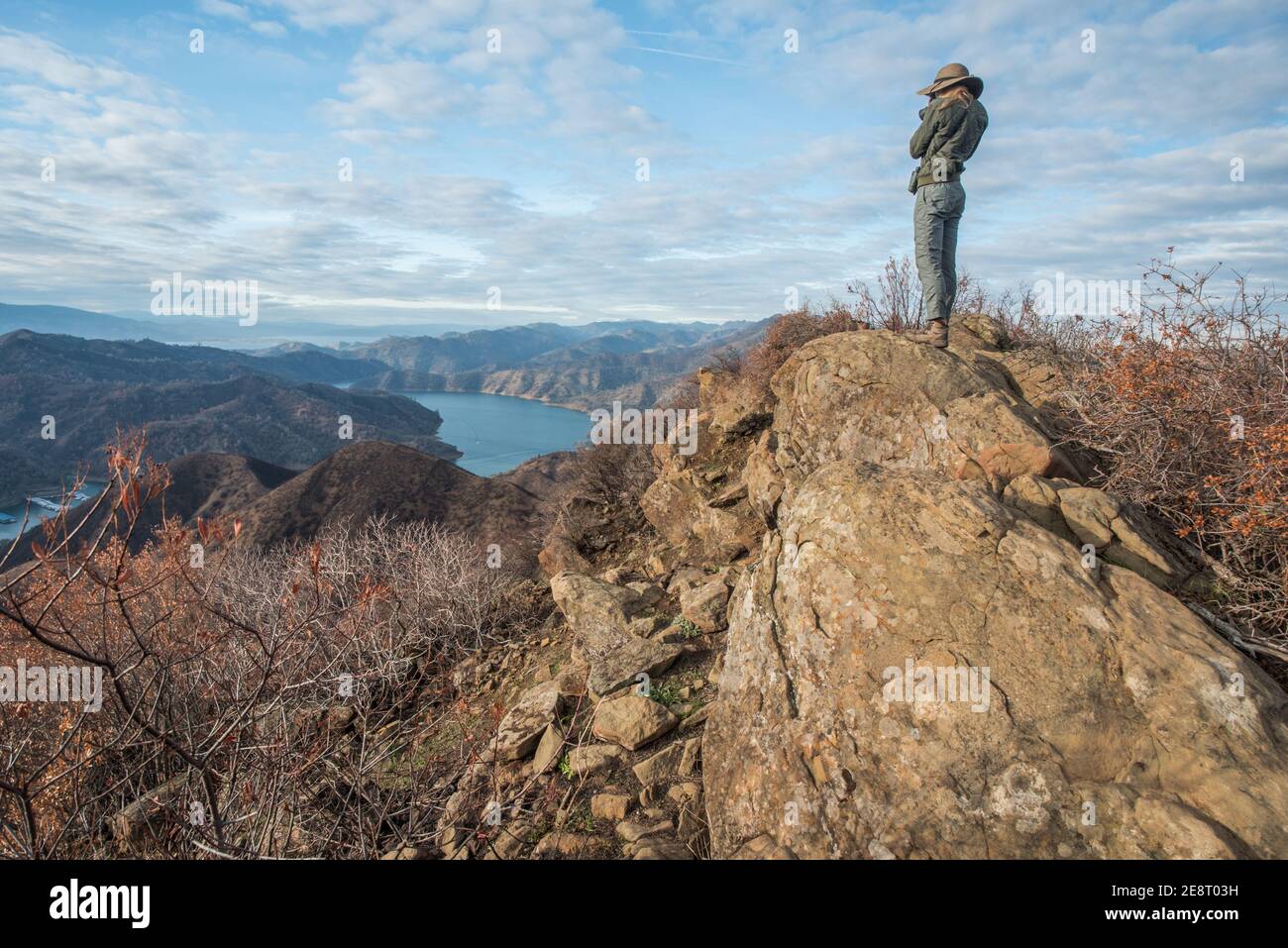 Una fit femminile escursionista si trova in cima a un grande masso che si affaccia sul paesaggio sottostante, tra cui un lago e le montagne. Foto Stock