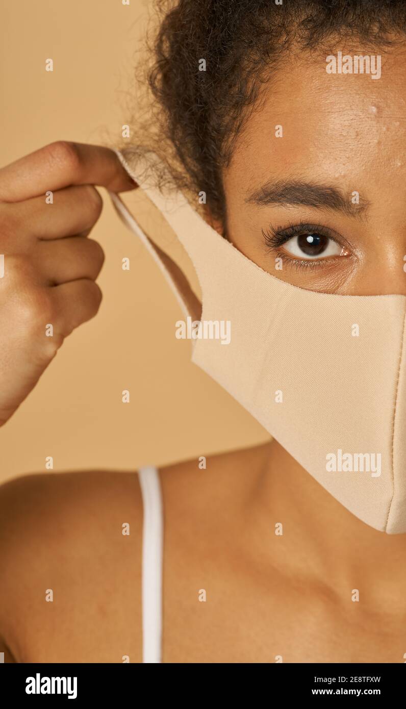 Ritratto a metà faccia di giovane donna di razza mista che regola la sua maschera facciale, guardando la fotocamera, in posa su sfondo beige. Sicurezza, concetto pandemico Foto Stock