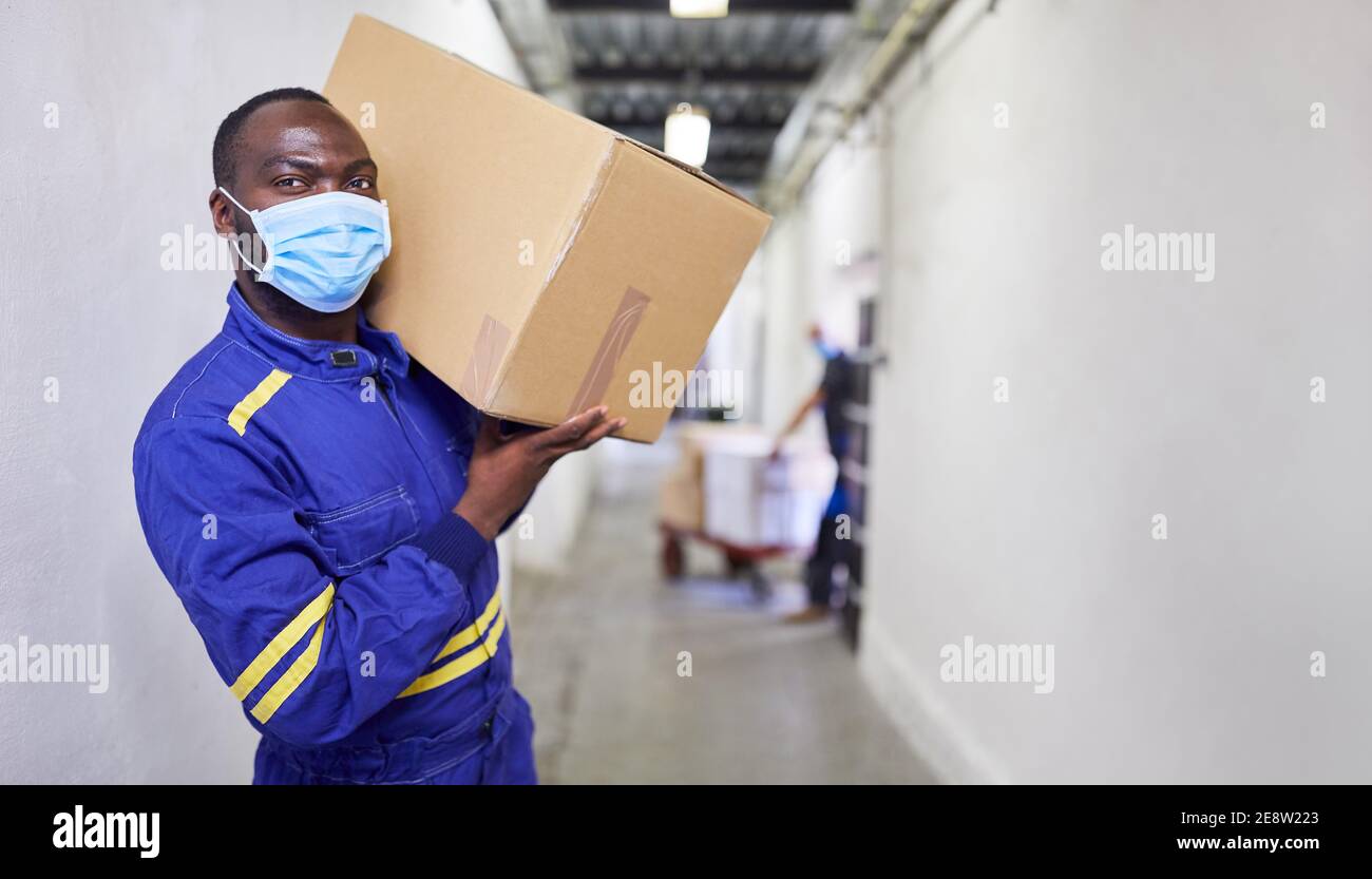 Operaio africano con maschera facciale a causa di Covid-19 porta un imballaggio nel magazzino Foto Stock