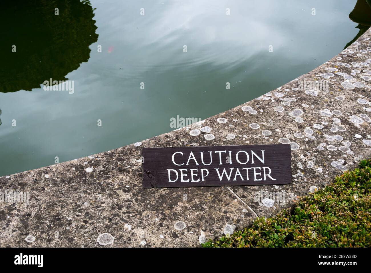 Attenzione segno di acqua profonda sul bordo coperto di lichen ad un laghetto ornamentale. Foto Stock