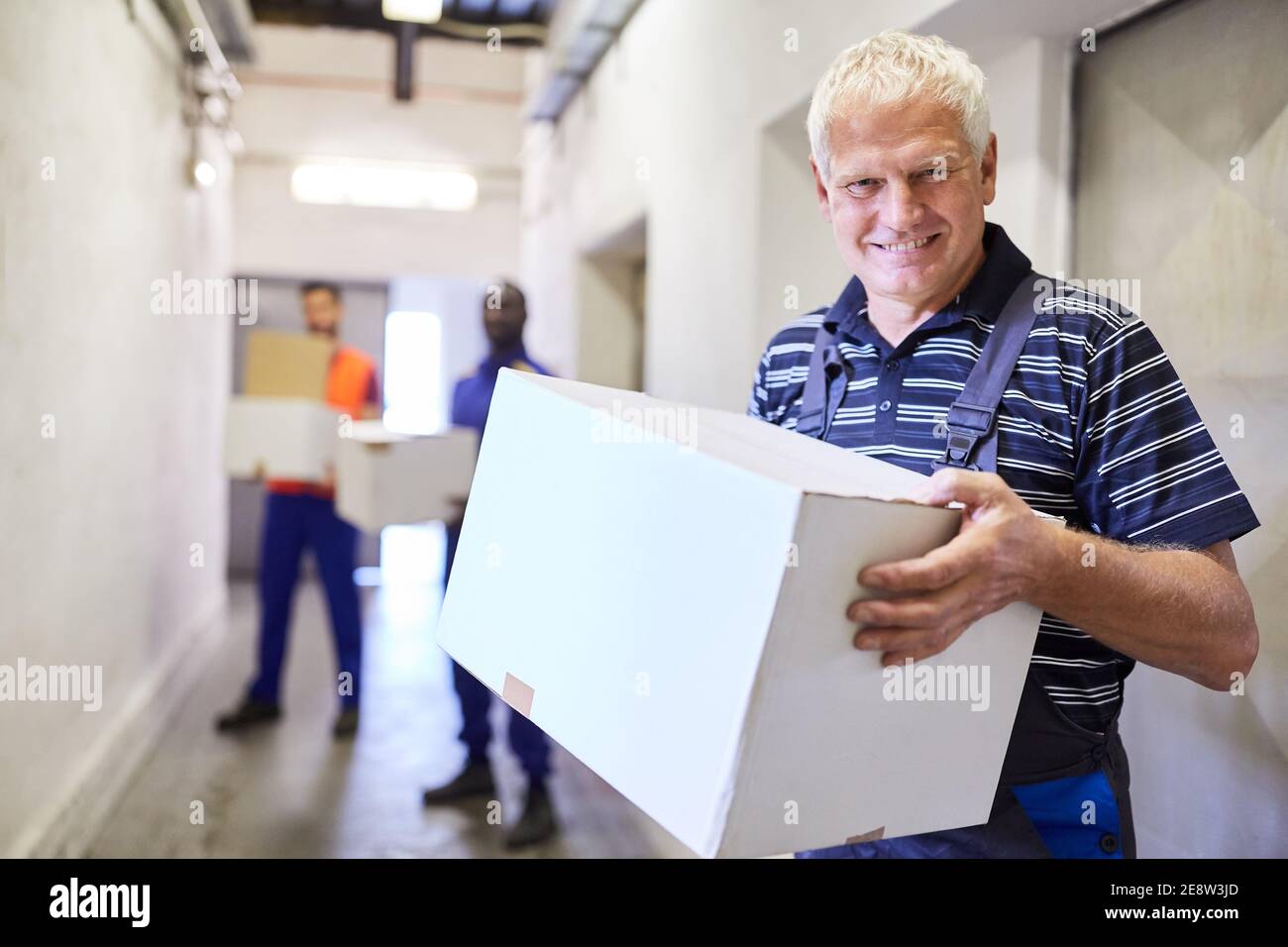 Il lavoratore sorridente come lavoratore temporaneo trasporta il pacchetto attraverso la corsia al magazzino di una fabbrica Foto Stock