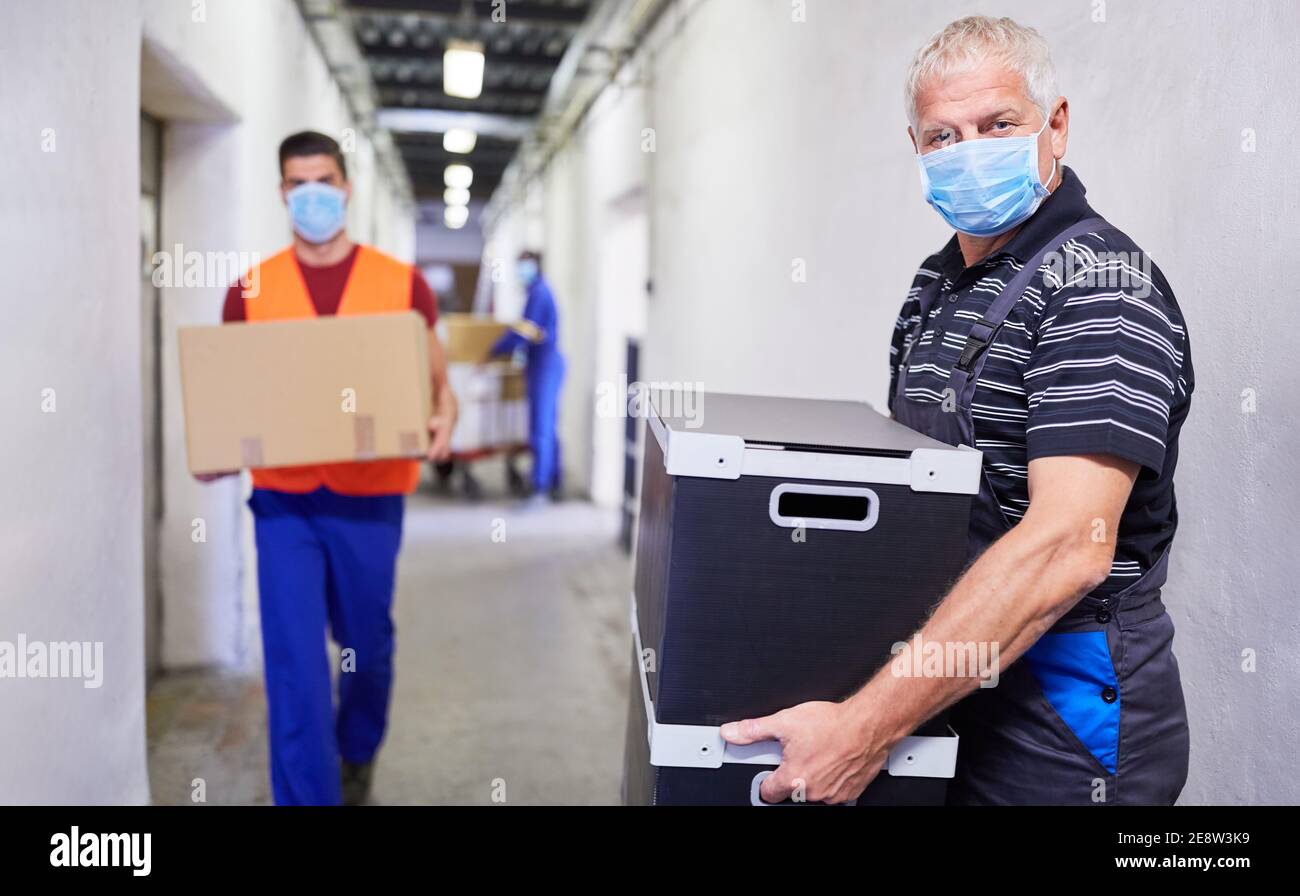 Il lavoratore più anziano con maschera facciale a causa di Covid-19 porta pacchetti nel magazzino di una fabbrica Foto Stock