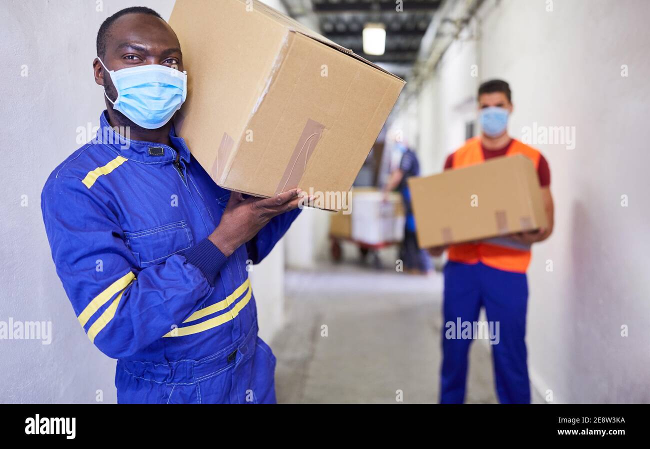 Due lavoratori con maschere per il viso a causa di Covid-19 trasportano cartone scatole in un magazzino Foto Stock