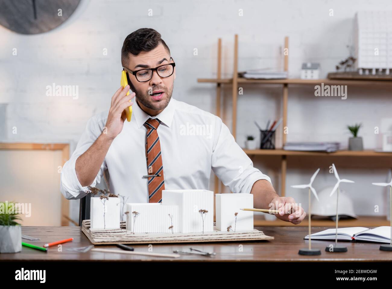 architetto che parla sullo smartphone sul posto di lavoro e punta con una matita a progetto architettonico Foto Stock