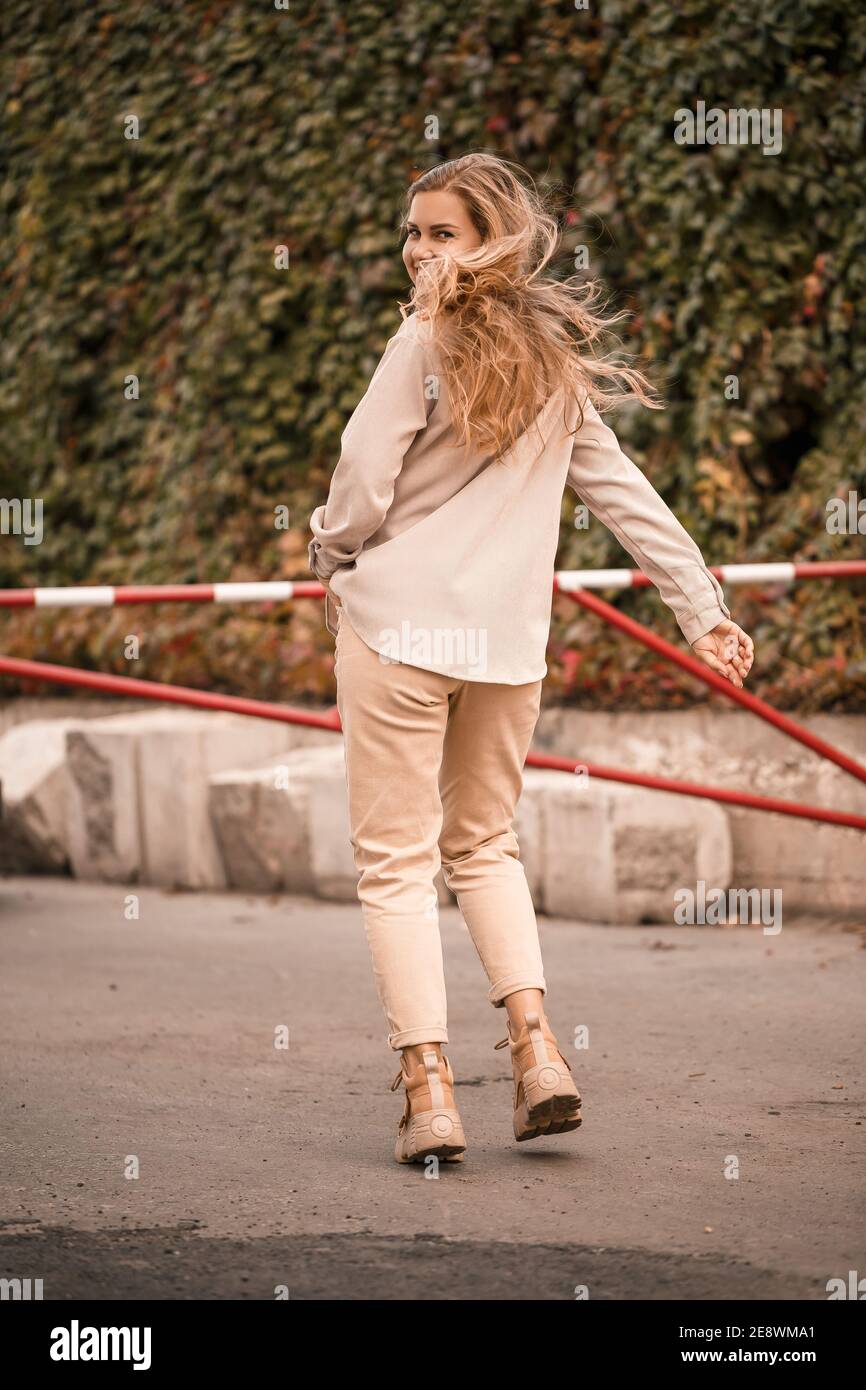 Una giovane bella bionda sta camminando per la strada, sta indossando jeans e una camicia beige. Bella ragazza vestita in stile casual con un sorriso sopra Foto Stock