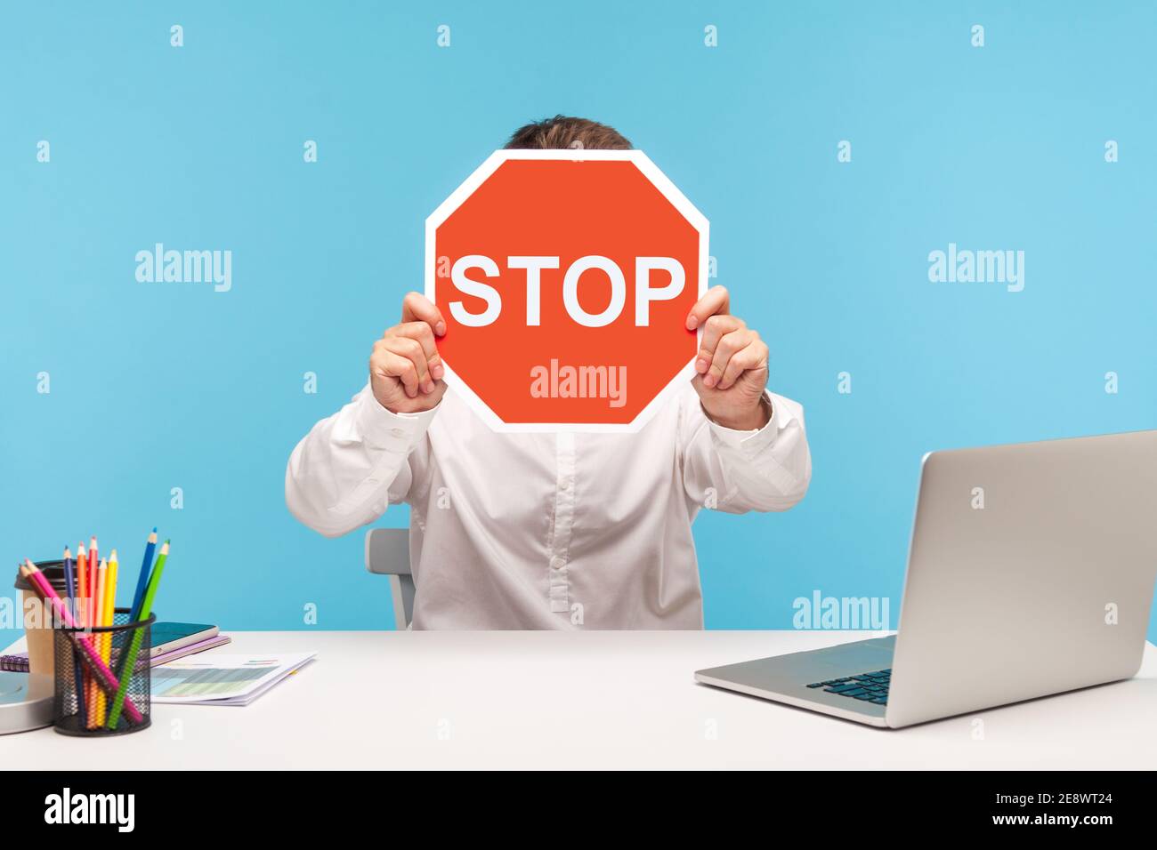 Uomo lavoratore in camicia bianca nascondendo il volto dietro il cartello rosso stop traffico, seduto sul posto di lavoro con il laptop, simbolo di restrizione di divieto. Interno Foto Stock