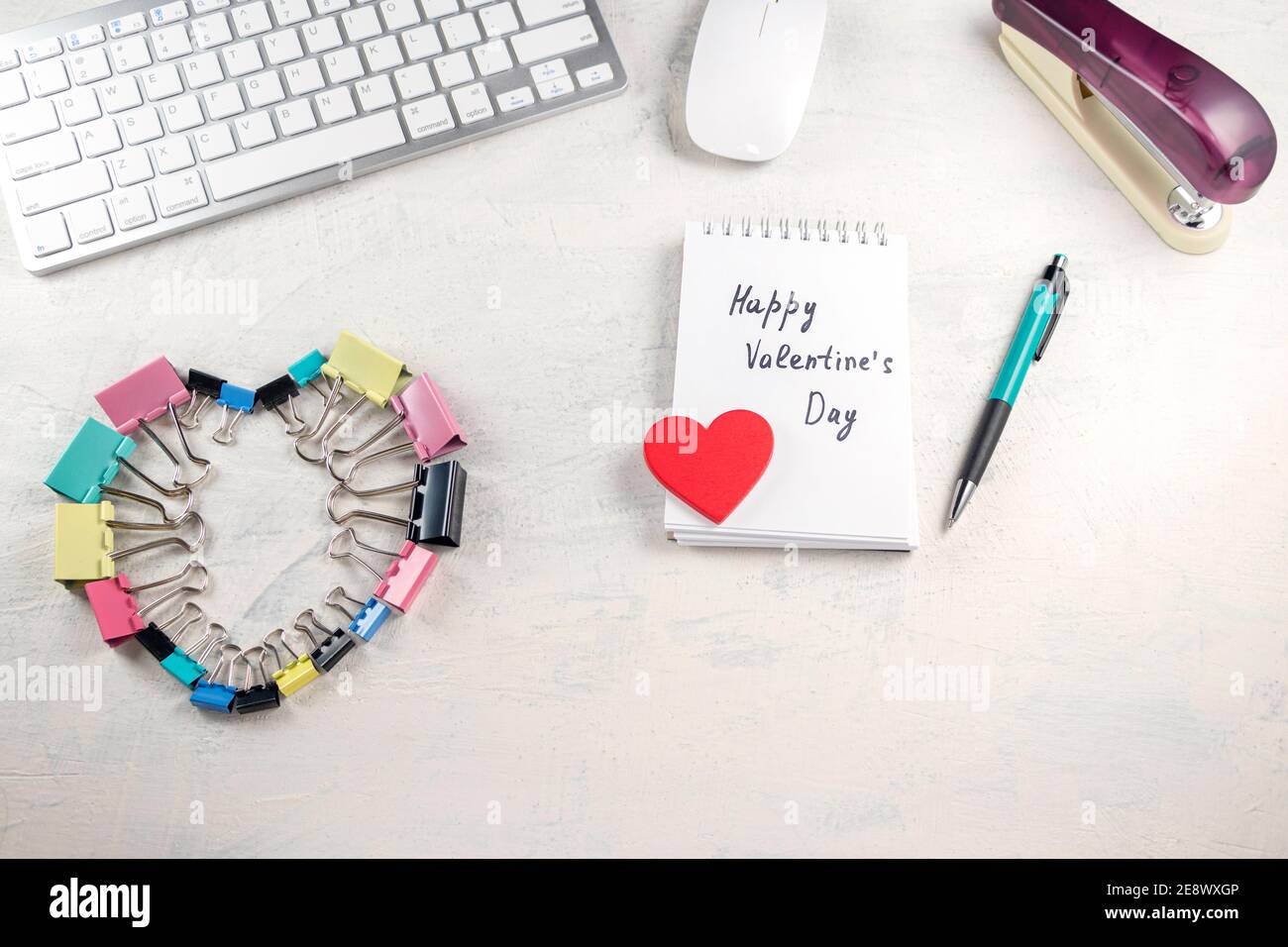 Le parole Happy Valentines giorno in una nota e cuore rosso sul desktop con clip cardio colorate. San Valentino in ufficio. Vista dall'alto Foto Stock