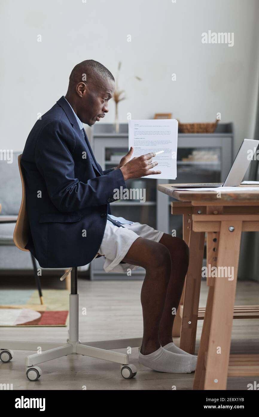Ritratto verticale con vista laterale di un uomo afro-americano con giacca formale e pantaloncini durante le riunioni online mentre si lavora da casa Foto Stock