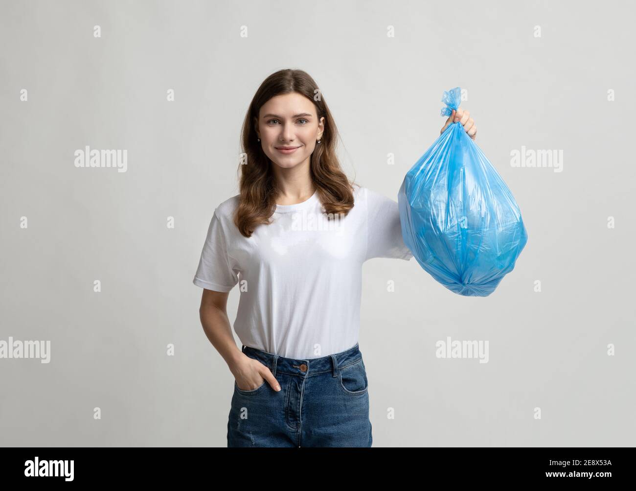 Felice giovane donna che tiene il sacchetto blu di plastica della spazzatura, in piedi su sfondo grigio Foto Stock