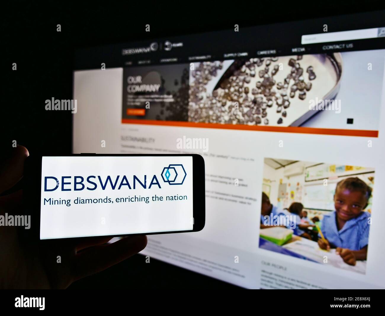 Persona che detiene il telefono cellulare con il logo della società mineraria Debswana Diamond Company Limited in esposizione davanti alla pagina web. Mettere a fuoco lo schermo del telefono. Foto Stock