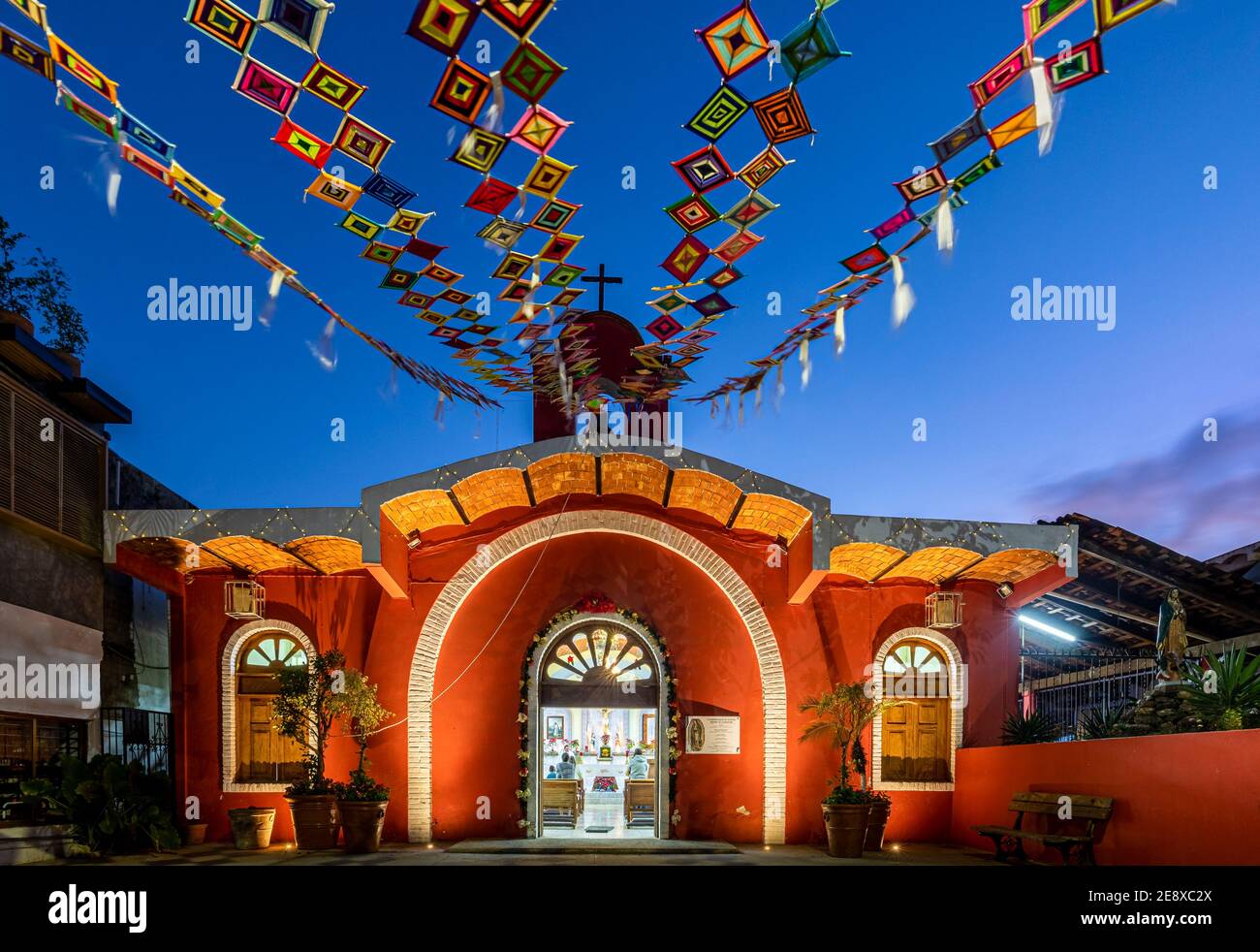 Piccola chiesa rossa sulla piazza con bandiere Huichol chiamata 'Ojos de Dios' che volano nel vento a Sayulita, Nayarit, Messico. Foto Stock