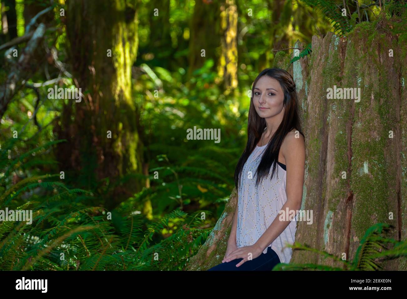 Una giovane donna con lunghi capelli marroni si appoggia contro un albero in un ambiente forestale. Foto Stock