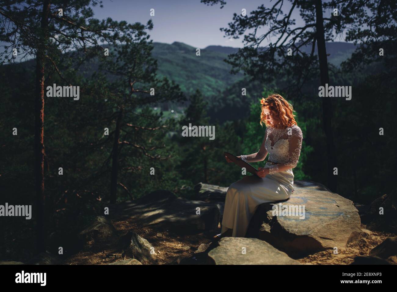 bella giovane sposa dai capelli rossi nella foresta con una corona floreale sulla testa. donna in lungo vestito bianco all'aperto il giorno d'estate. giorno di nozze. Foto Stock