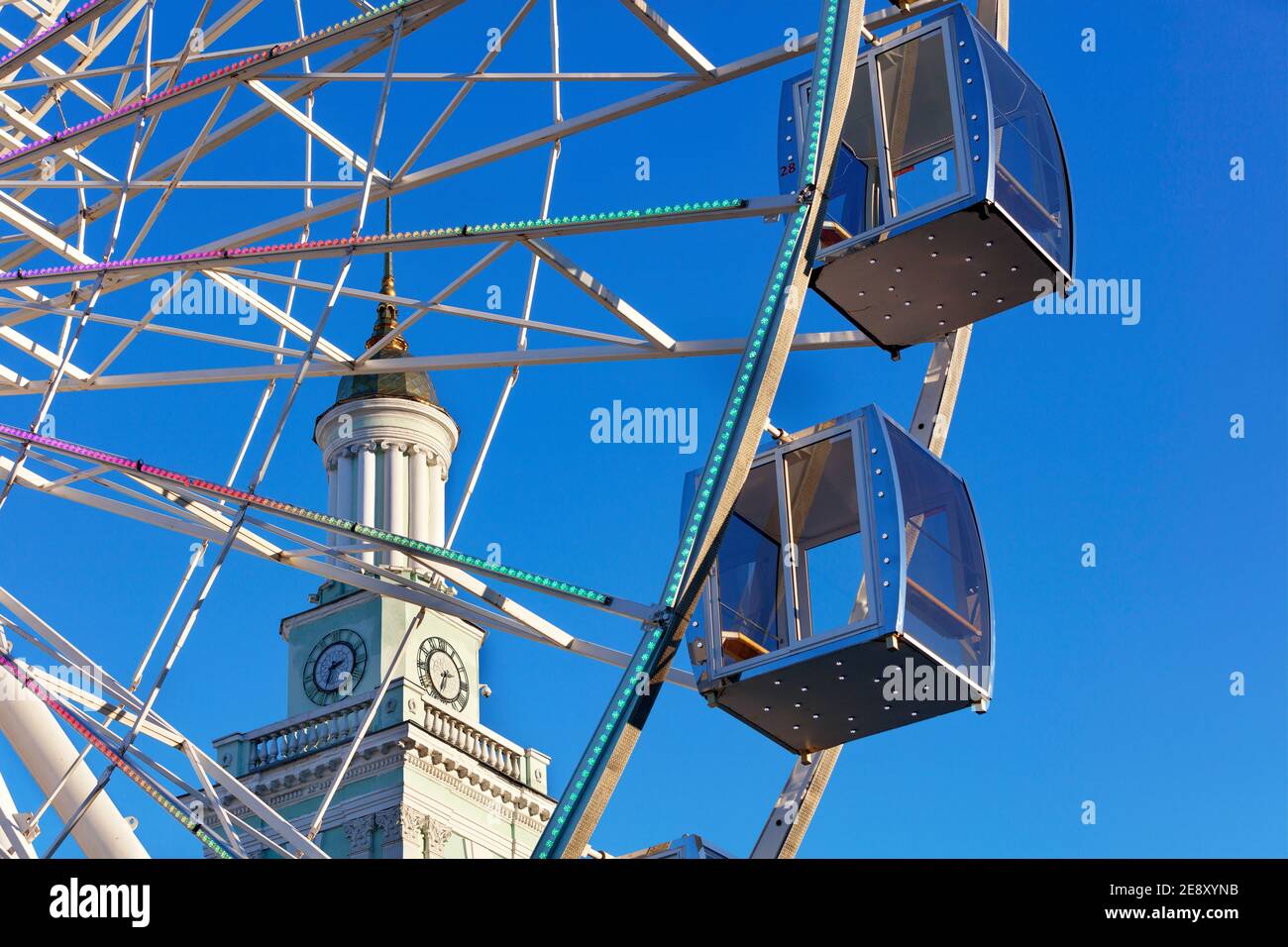 Una bella e panoramica vista delle cabine vuote della ruota panoramica contro il cielo blu e la guglia della vecchia torre dell'orologio nel pomeriggio. Foto Stock