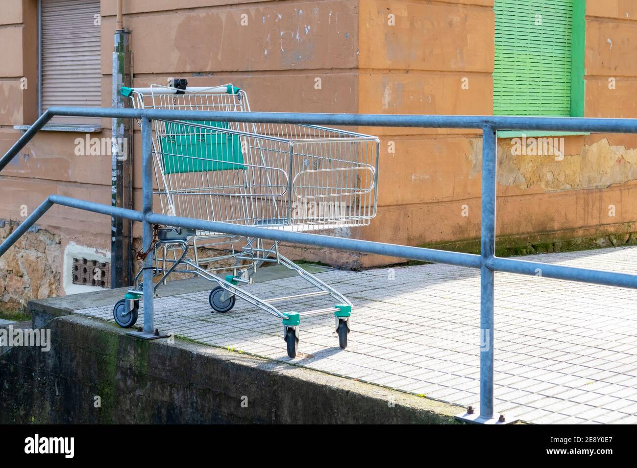 Abbandonato carrello di alimentari vuoto in un vecchio cortile di strada con pareti in shabby e persiane chiuse. Carrello della spesa scartato incatenato ai corrimano di un c Foto Stock