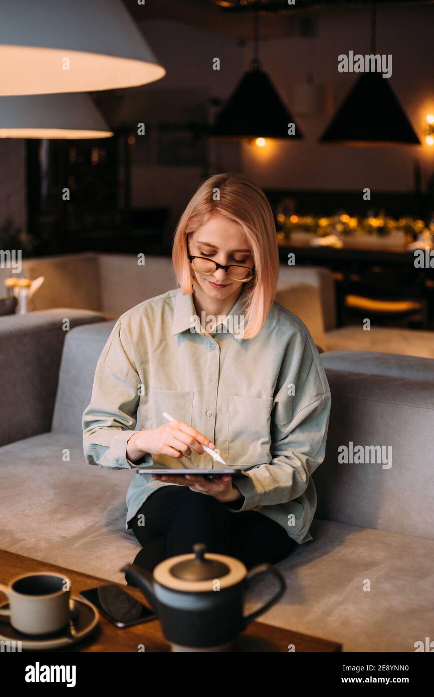 Giovane designer creativa donna in occhiali che tengono stilo disegno penna su tablet digitale mentre si siede in un moderno cafe Foto Stock