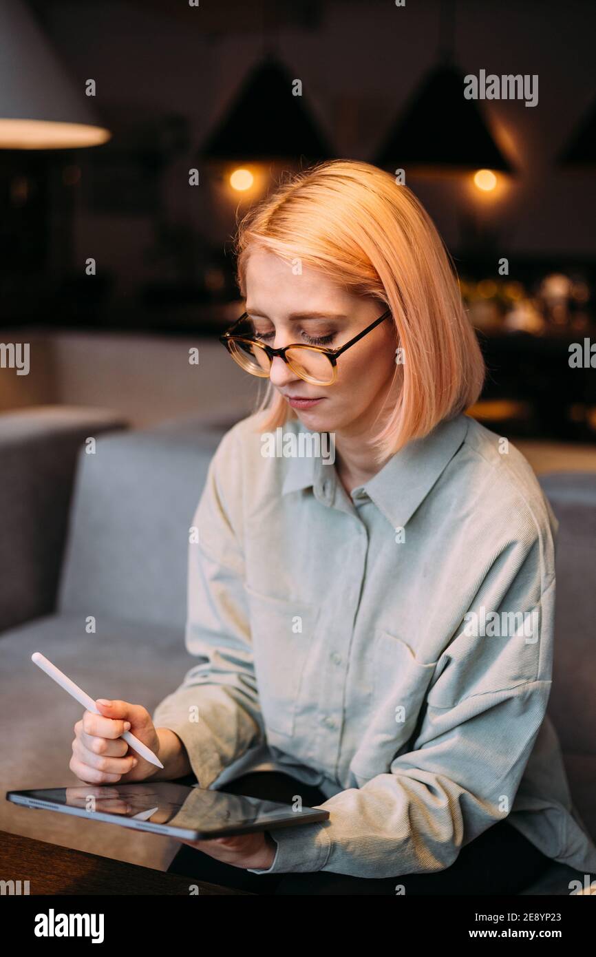 Giovane designer creativa donna in occhiali che tengono stilo disegno penna su tablet digitale mentre si siede in un moderno cafe Foto Stock