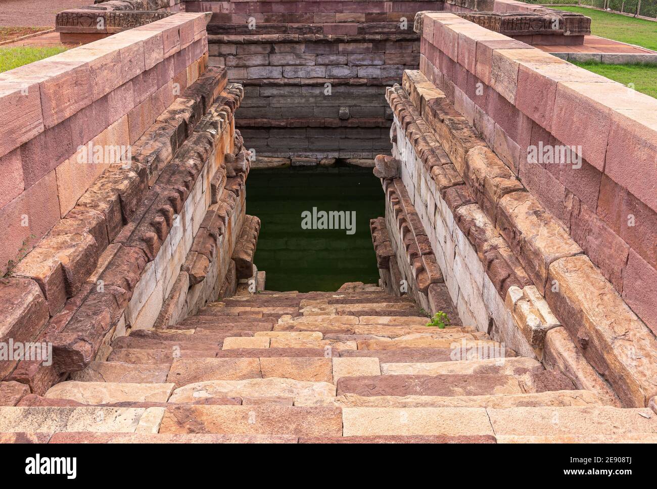 Aihole, Karnataka, India - 7 novembre 2013: Huchchimalli Gudi o Tempio. Scalinata in giù nel serbatoio d'acqua verde fiancheggiata da pareti di pietra rossastre e marroni Foto Stock