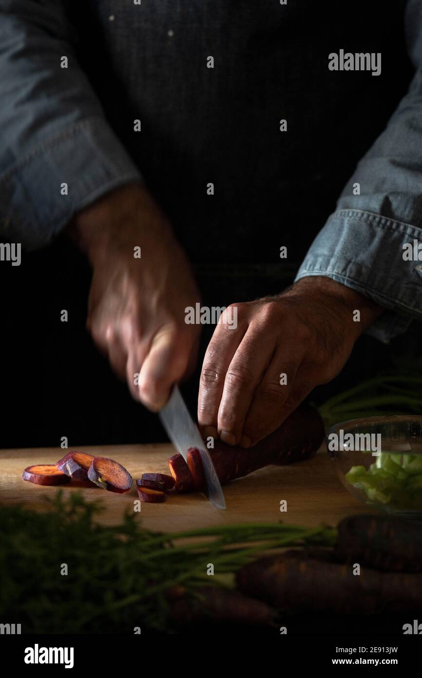 Un primo piano di uno chef che trita una carota viola su un tagliere di legno. Foto Stock