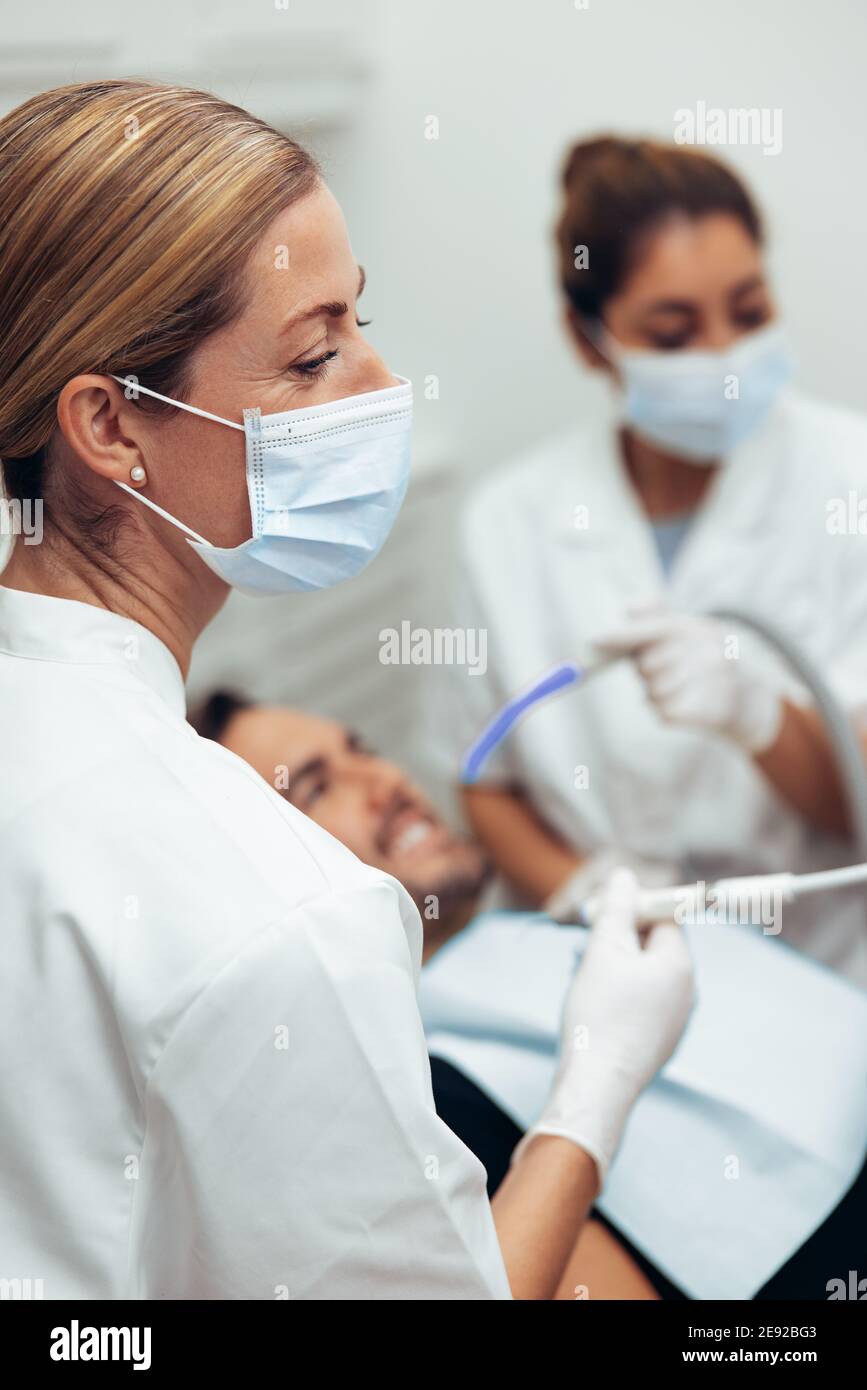 Primo piano di una chirurgo dentale femminile che tratta un paziente in clinica con il suo assistente in background. Dentista che tratta un paziente di sesso maschile nel suo ufficio. Foto Stock