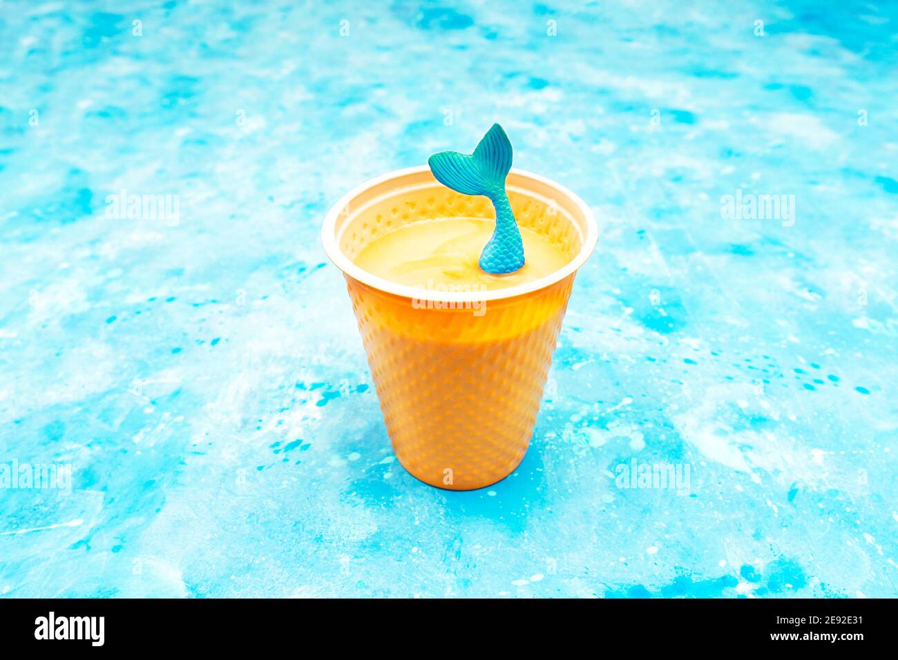 La coda della sirena giocattolo fuoriesce da una tazza di plastica di cappuccino su sfondo azzurro Foto Stock