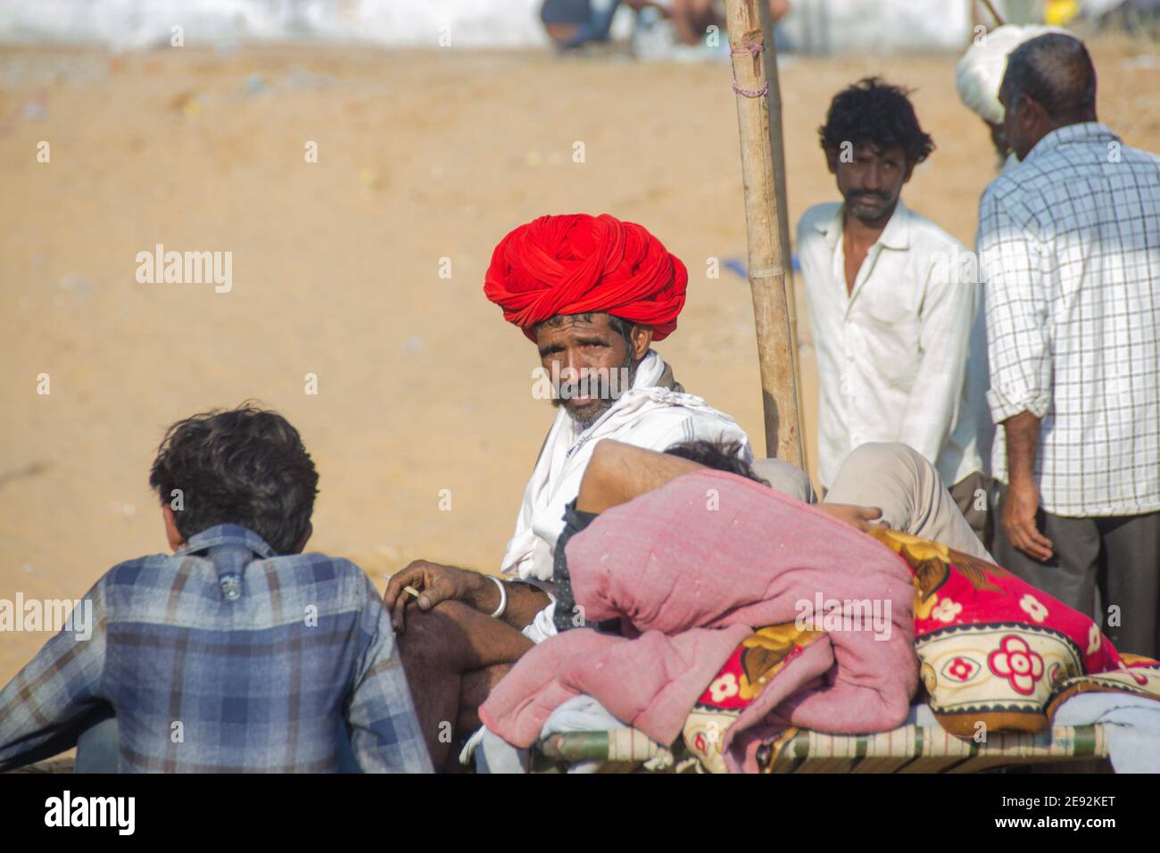 un gestore di cammelli è seduto sui terreni fieristici al sole del mattino. Il turbante rosso sulla sua testa è luminoso e attraente. Foto Stock