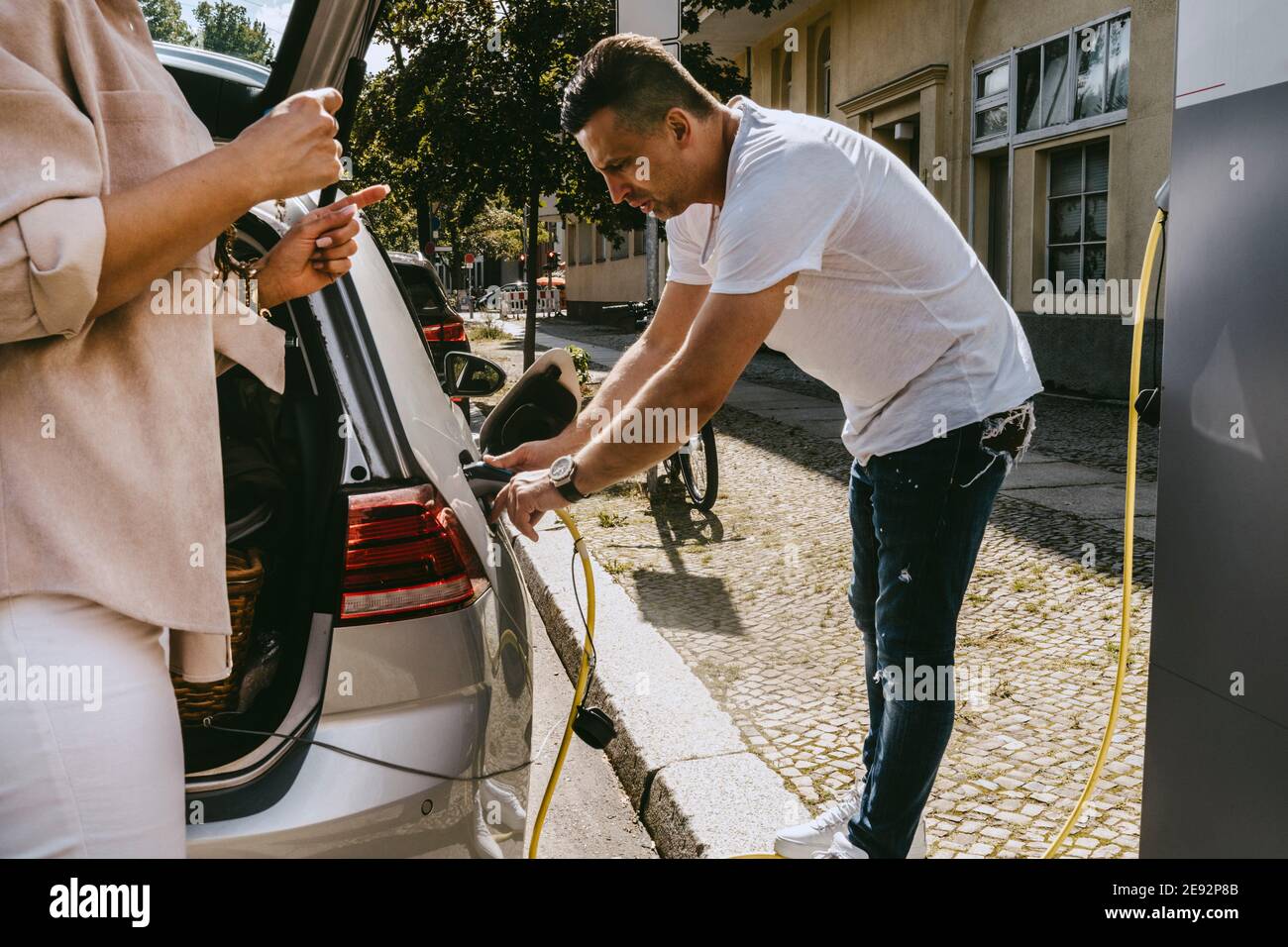 Padre che carica auto elettrica mentre madre in piedi vicino a bordo strada Foto Stock