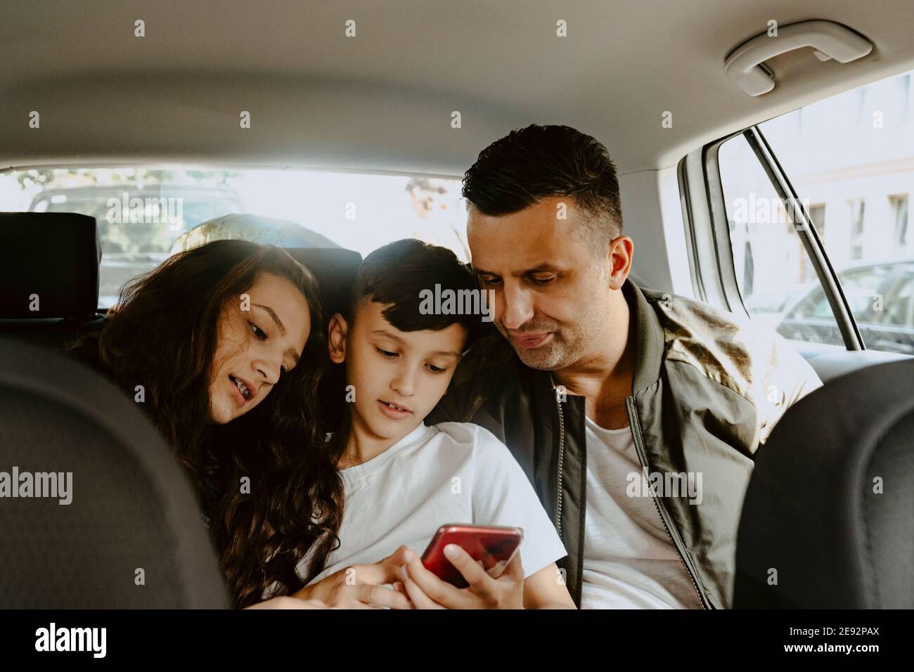 Ragazzo che usa il telefono cellulare seduto con padre e sorella dentro auto elettrica Foto Stock