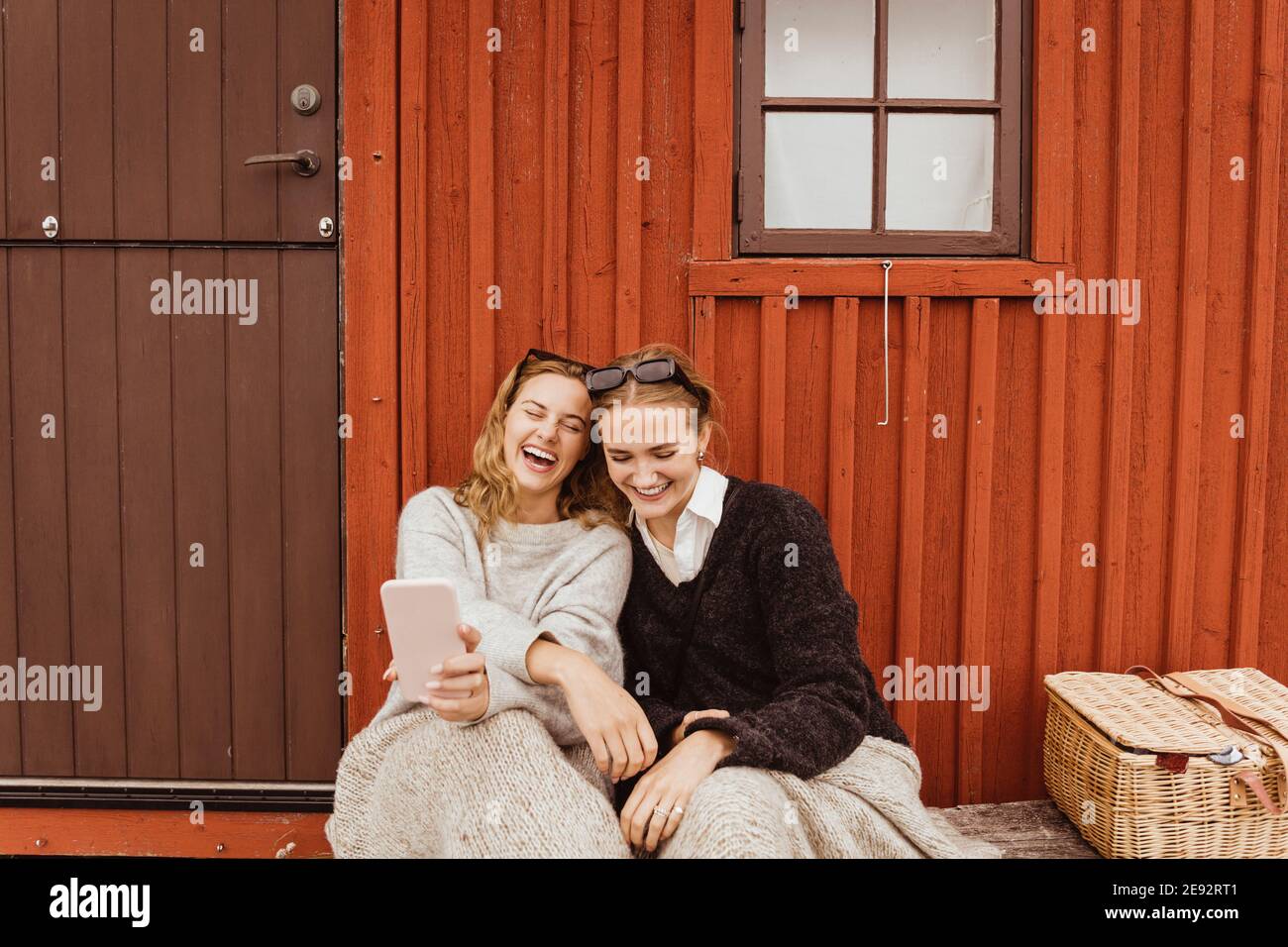 Le amiche ridono mentre prendono selfie sul telefono cellulare contro cottage Foto Stock