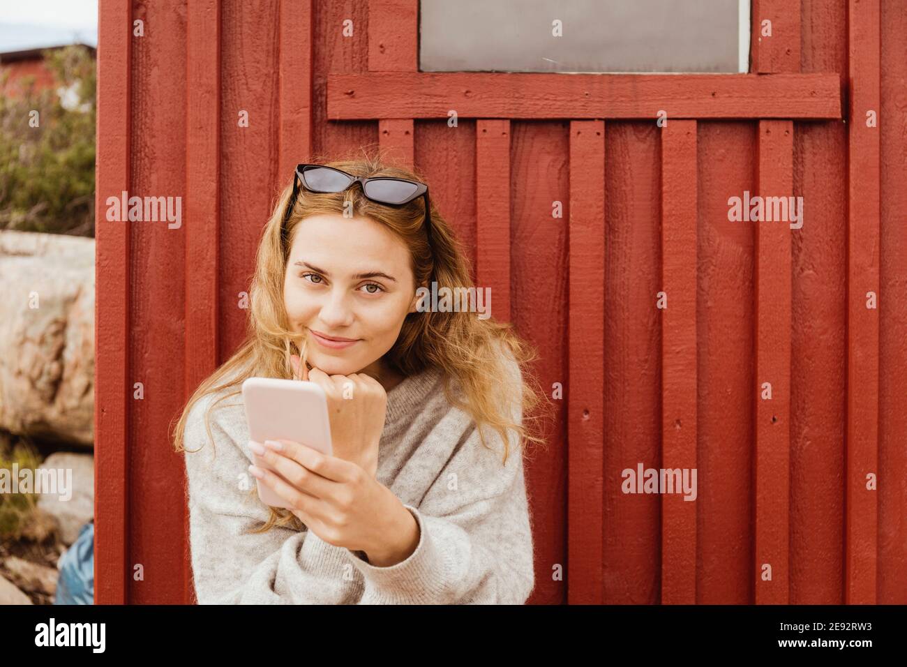 Donna sorridente con la mano sul mento che tiene lo smartphone contro cottage Foto Stock