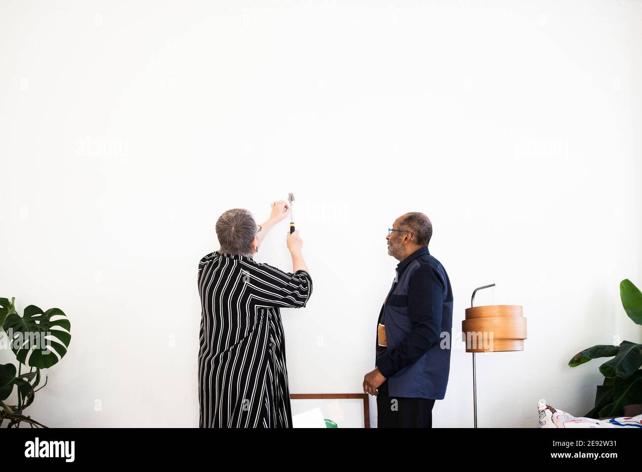 Donna anziana martellando chiodo su parete per fissare la pittura in piedi da un uomo anziano in soggiorno Foto Stock
