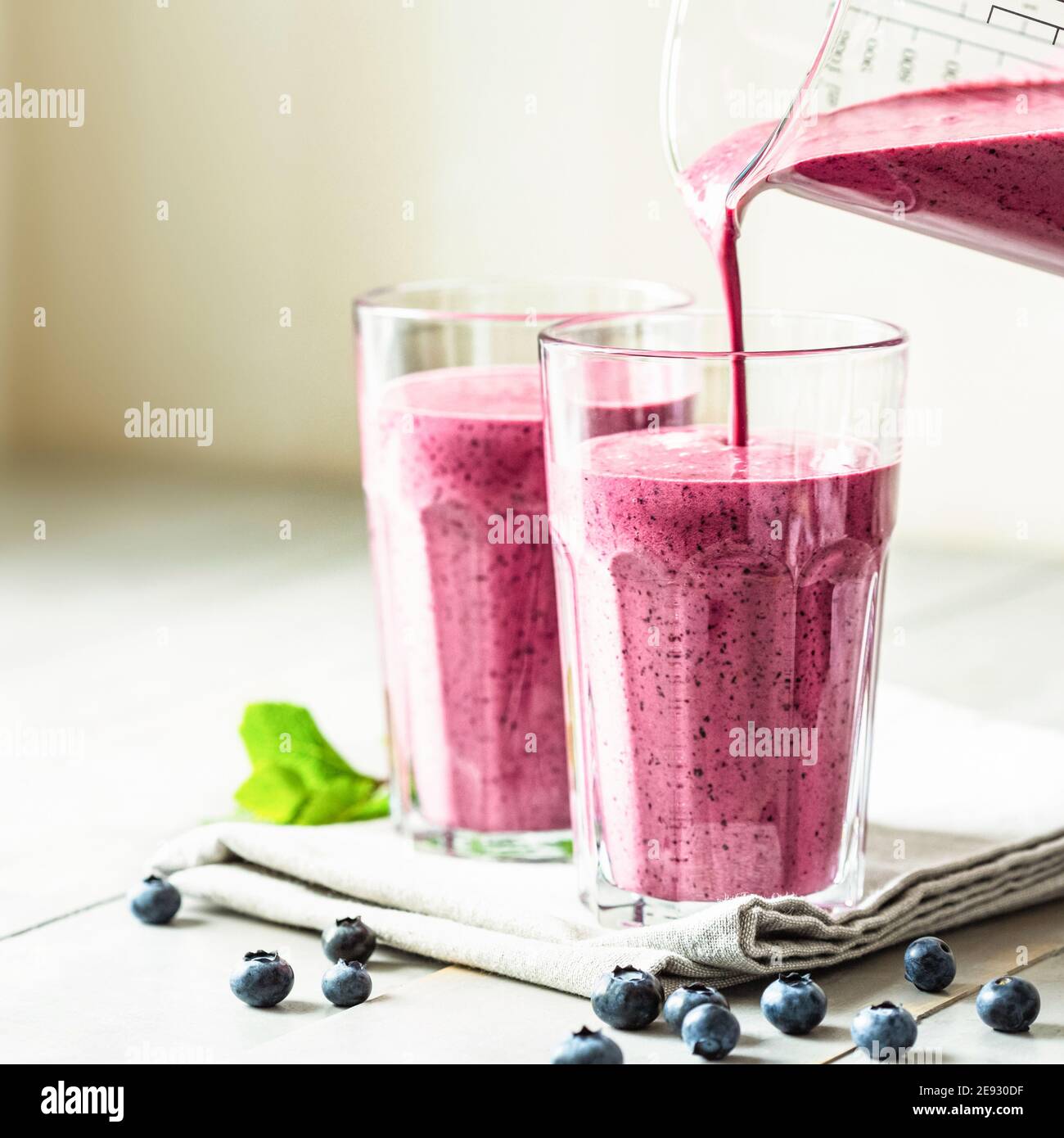 Due bicchieri di frullato al mirtillo con guarnitura alla menta e paglia sul tavolo. Il bacca frullato viene versato in un bicchiere. Foto Stock