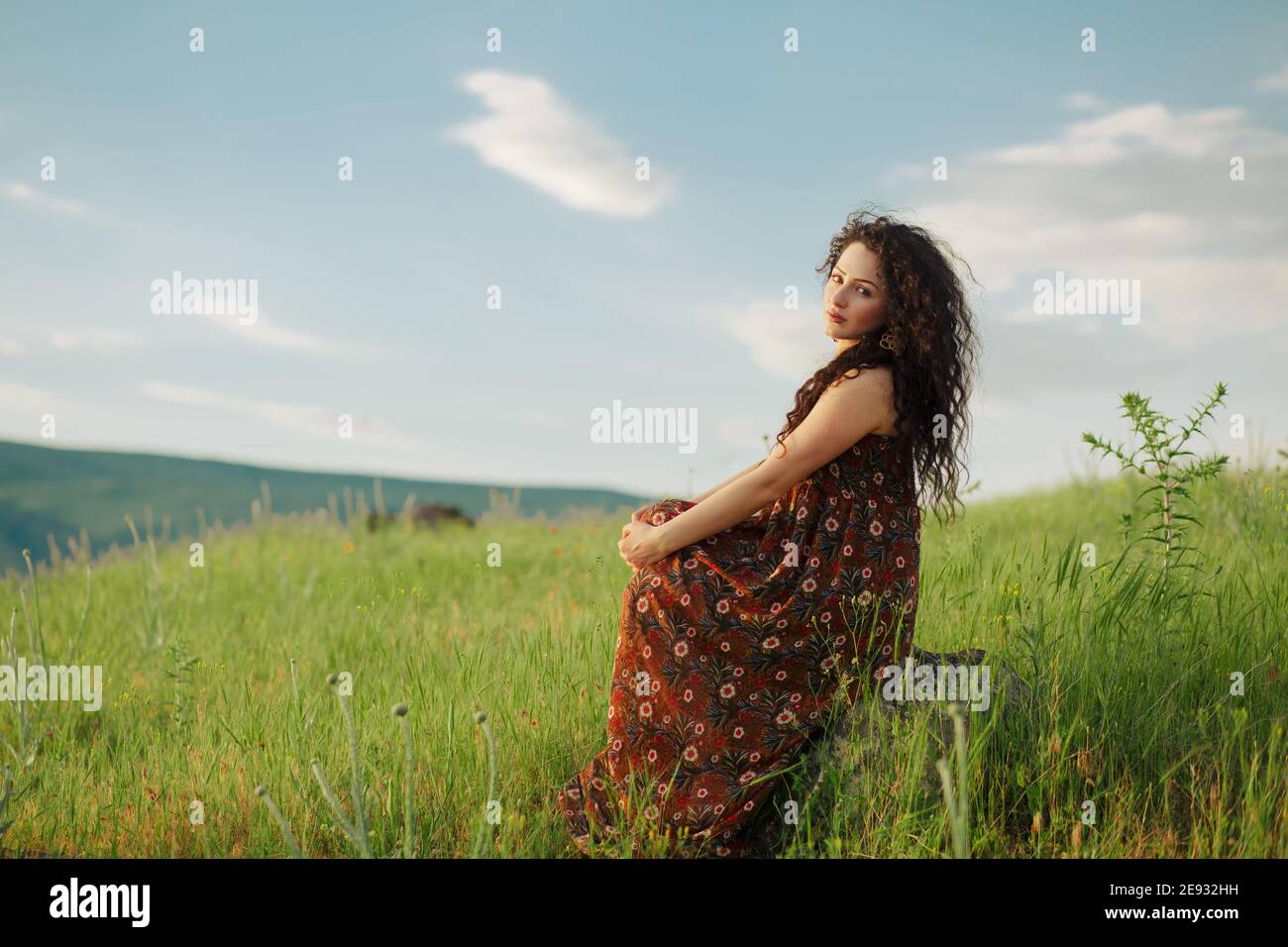 Stile di vita scatto di capelli ricci ragazza seduta su pietra in campo e guardando la fotocamera Foto Stock
