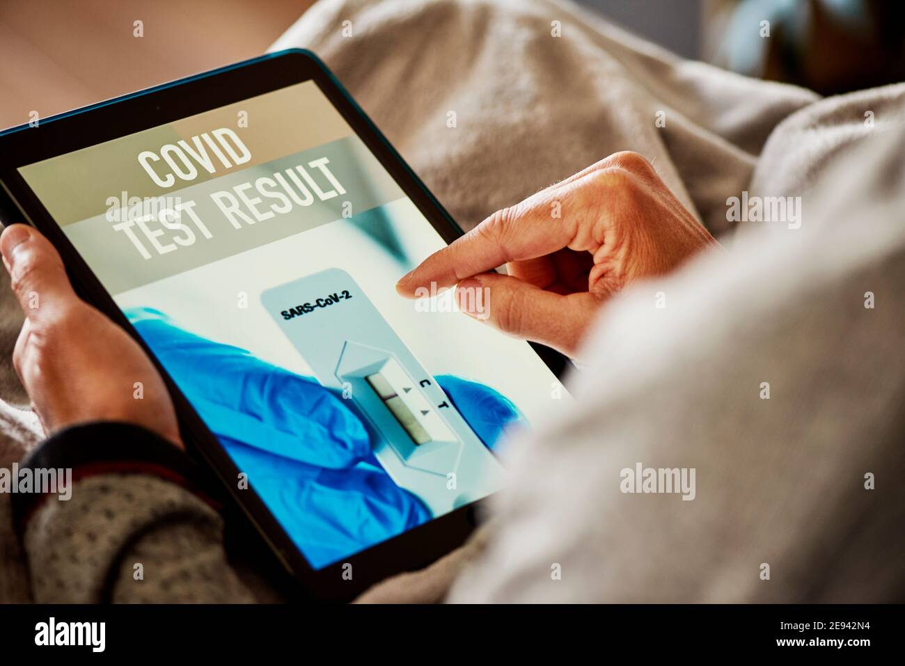 primo piano di un giovane caucasico in pigiama, seduto su un divano avvolto in una coperta grigia, controllando il risultato del suo covid test in un'app simulata in h Foto Stock