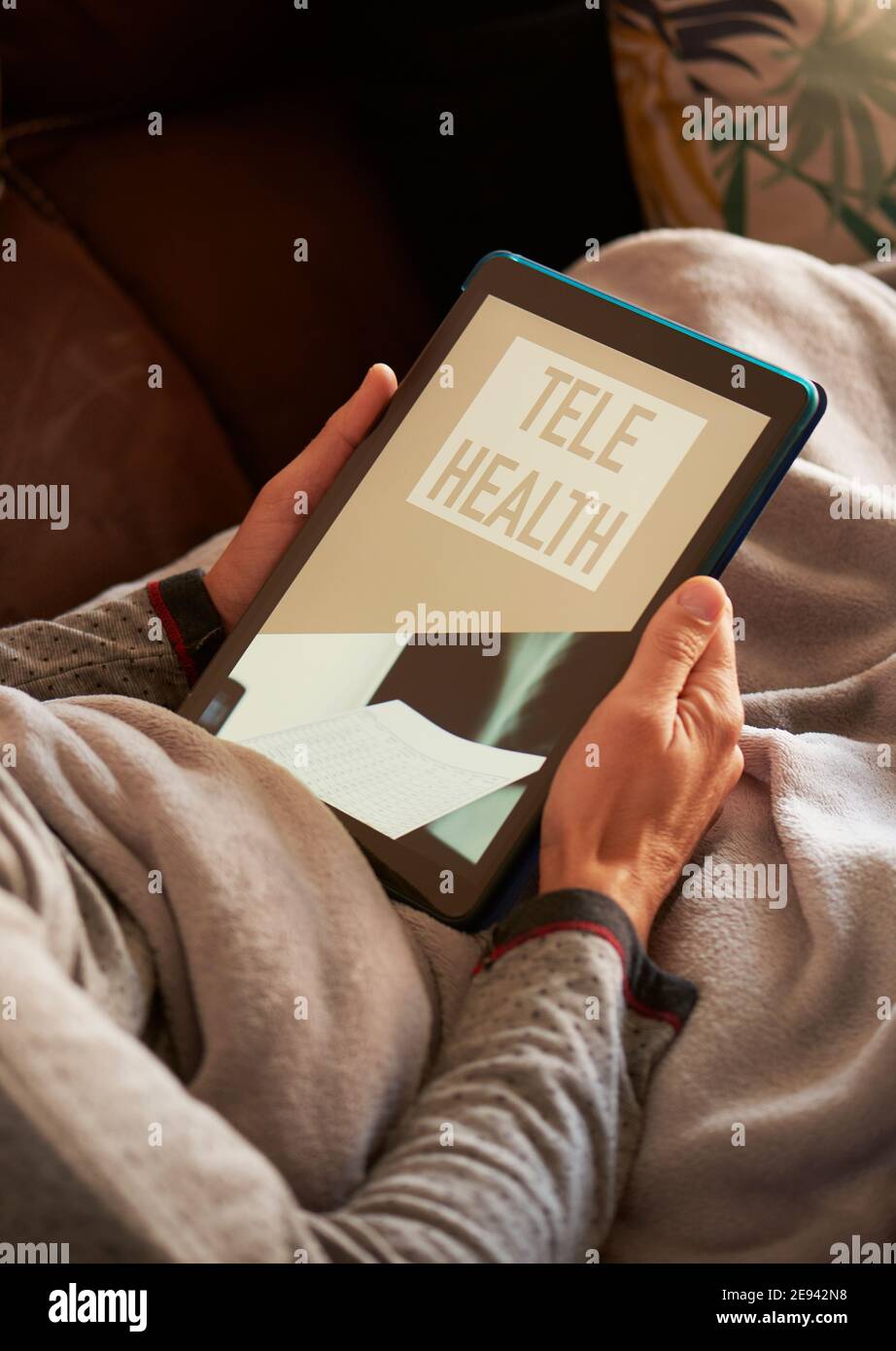 un giovane uomo caucasico, comodamente sdraiato in un divano avvolto in una coperta grigia calda, ha la sua tavoletta in mano con il testo teleghealth sulla sua scra Foto Stock
